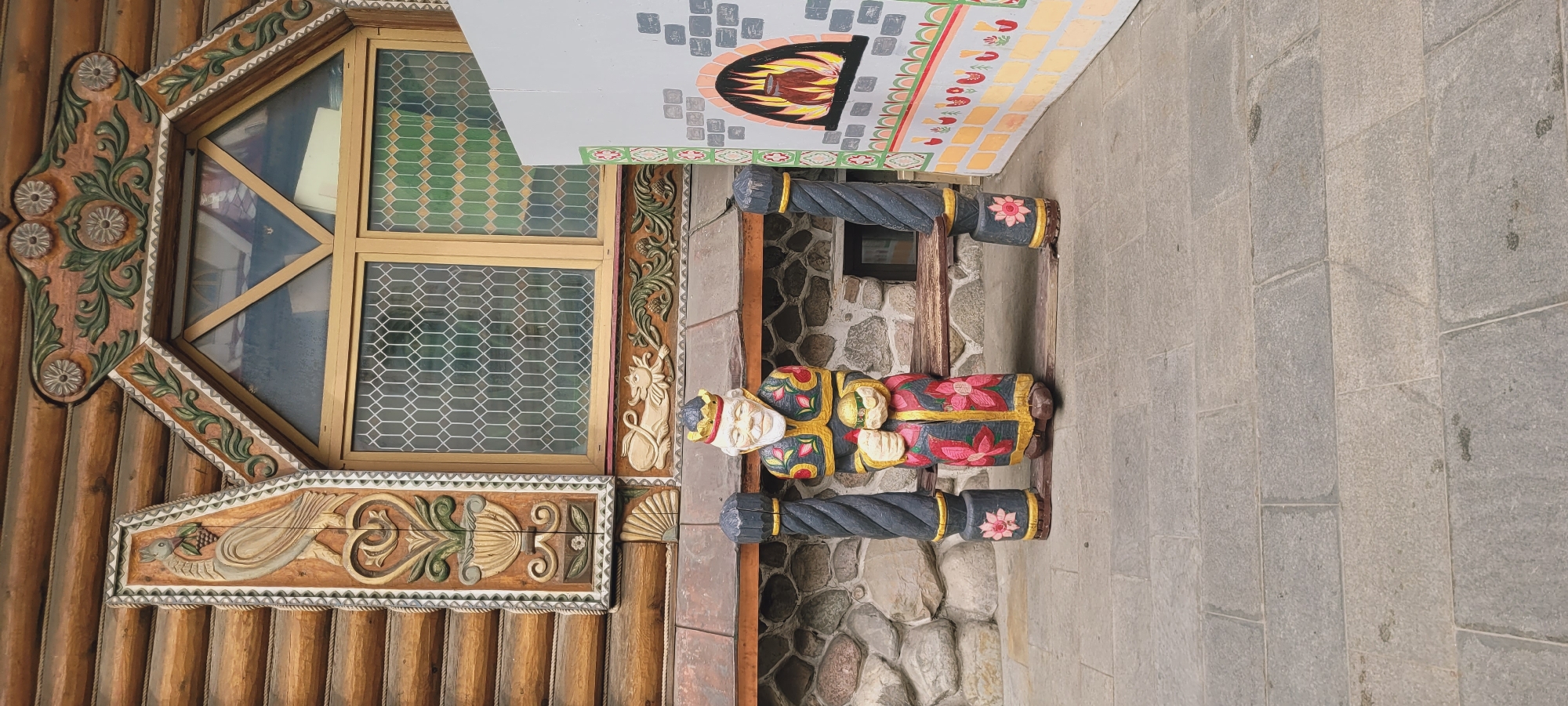 The image shows a section of a building with intricate wooden carvings and a window with a geometric pattern. The building is made of stacked wooden logs. The window frame is decorated with carved flowers and geometric patterns. There's a smaller section with a carving of a bird and decorative swirls. To the right of the building is a stone wall. In front of the wall is a carved figure of a person. Above the figure is a sign with an image of a fireplace. The ground is paved with rectangular stone blocks. The location is not specified in the prompt.