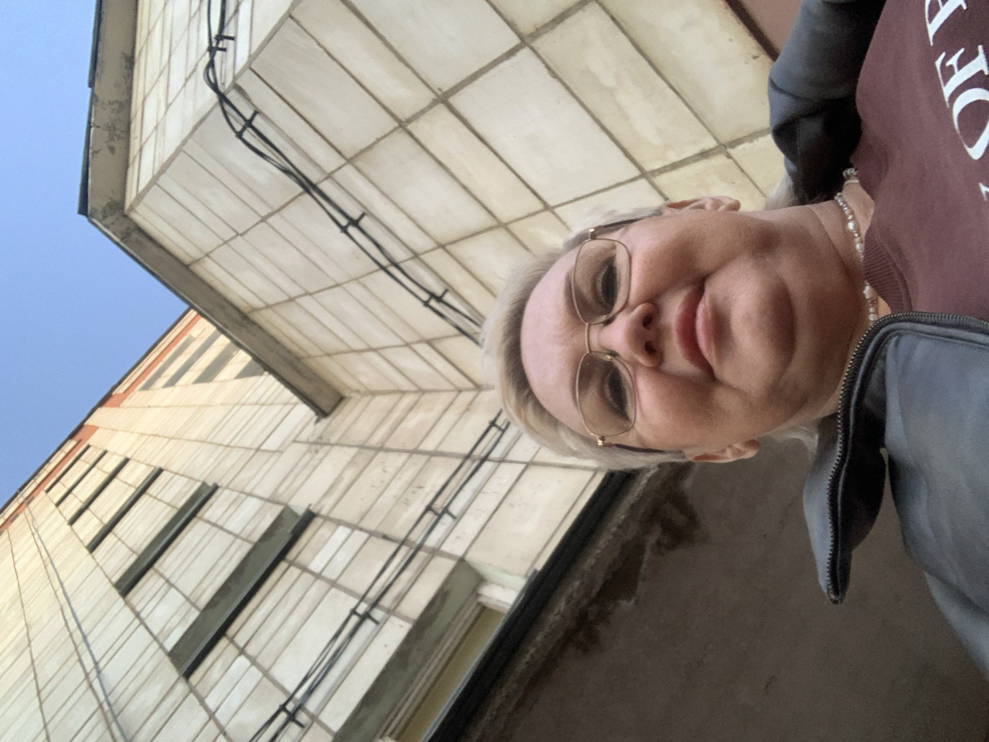 A woman with blonde hair, wearing glasses, a maroon sweatshirt with white writing, and a black leather jacket, takes a low-angle selfie. She is looking up at a tall, light-colored building with a grid-like pattern of rectangular tiles and windows. Black wires are visible running along the building's facade. The sky is blue in the upper left corner. Based on the context, this is likely in Chaikovsky, Russia, as that information was provided and there are no indications of a different location.