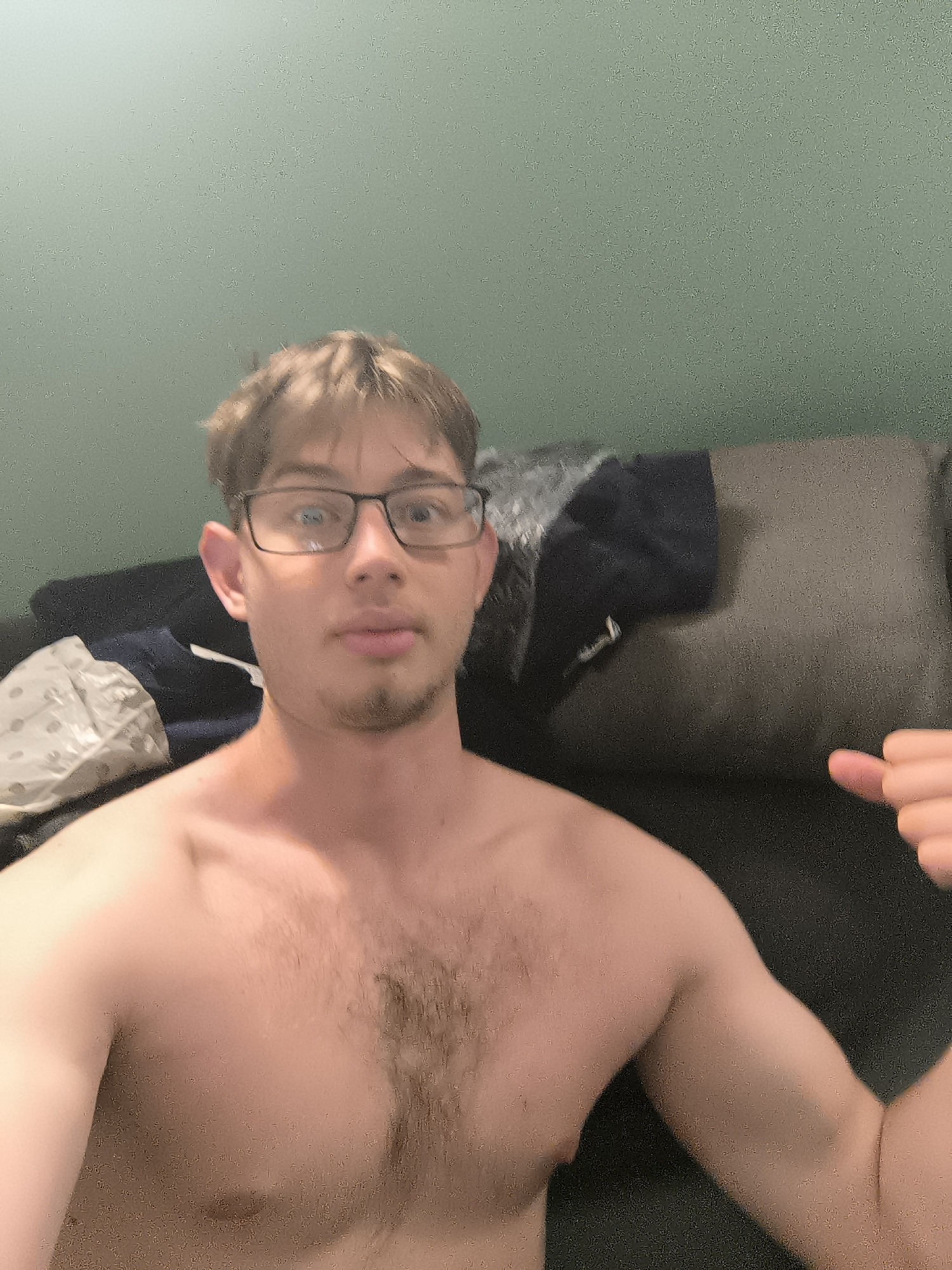A shirtless man with glasses sits on a dark-colored couch in an indoor setting. The background is a plain green wall. Other items, including what appears to be clothing, are visible on the couch beside him. The image does not show any indicators pointing to Sofia, Bulgaria.