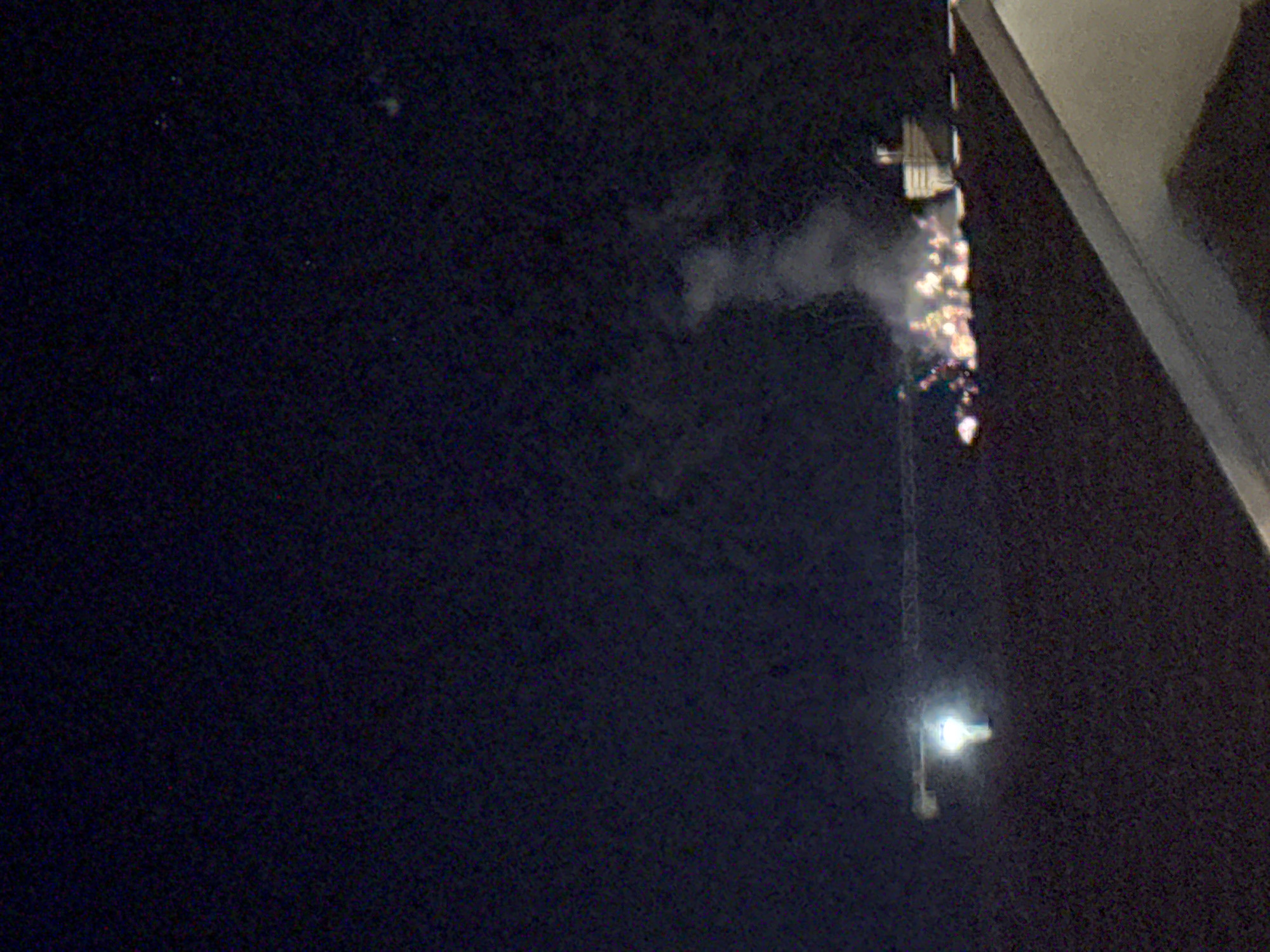 The image captures a nighttime scene with a dark, starry sky. In the mid-ground, a construction crane is visible with a bright light illuminating its structure. To the right of the crane, there appears to be a structure with lights and what looks like smoke or steam rising from it. A line of lights, possibly from a building or event, extends horizontally across the scene. The foreground shows the edge of a light-colored surface, likely a rooftop or a balcony, obscuring the lower part of the image. The scene suggests an urban or industrial setting at night, possibly in the area of Trap, Montenegro.