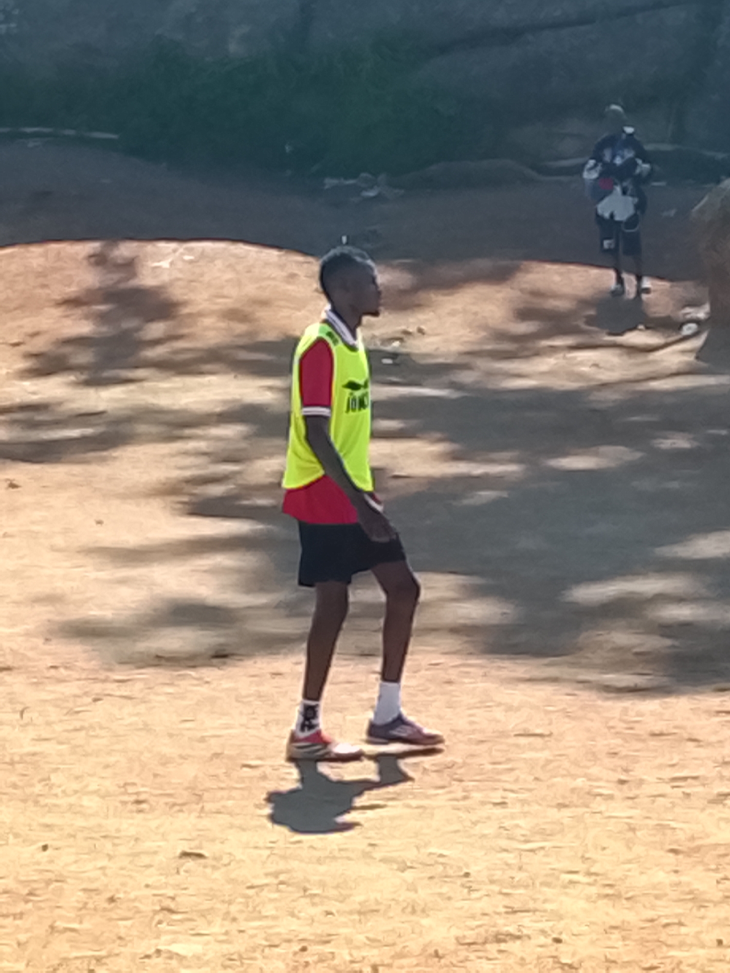 A young Black male, appearing to be a teenager, stands on a dirt field in what is likely a park or recreational area in Jos, Nigeria. He is wearing a yellow soccer training vest over a red shirt with white trim on the sleeves, and black shorts. He is also wearing white socks and athletic shoes. The ground is a dusty, sandy color, with scattered shadows suggesting it is daytime and sunny. In the background, another person, also appearing to be Black and male, is visible wearing a patterned jersey and shorts, possibly also engaged in athletic activity. The environment appears somewhat rustic, with natural terrain and sparse vegetation.