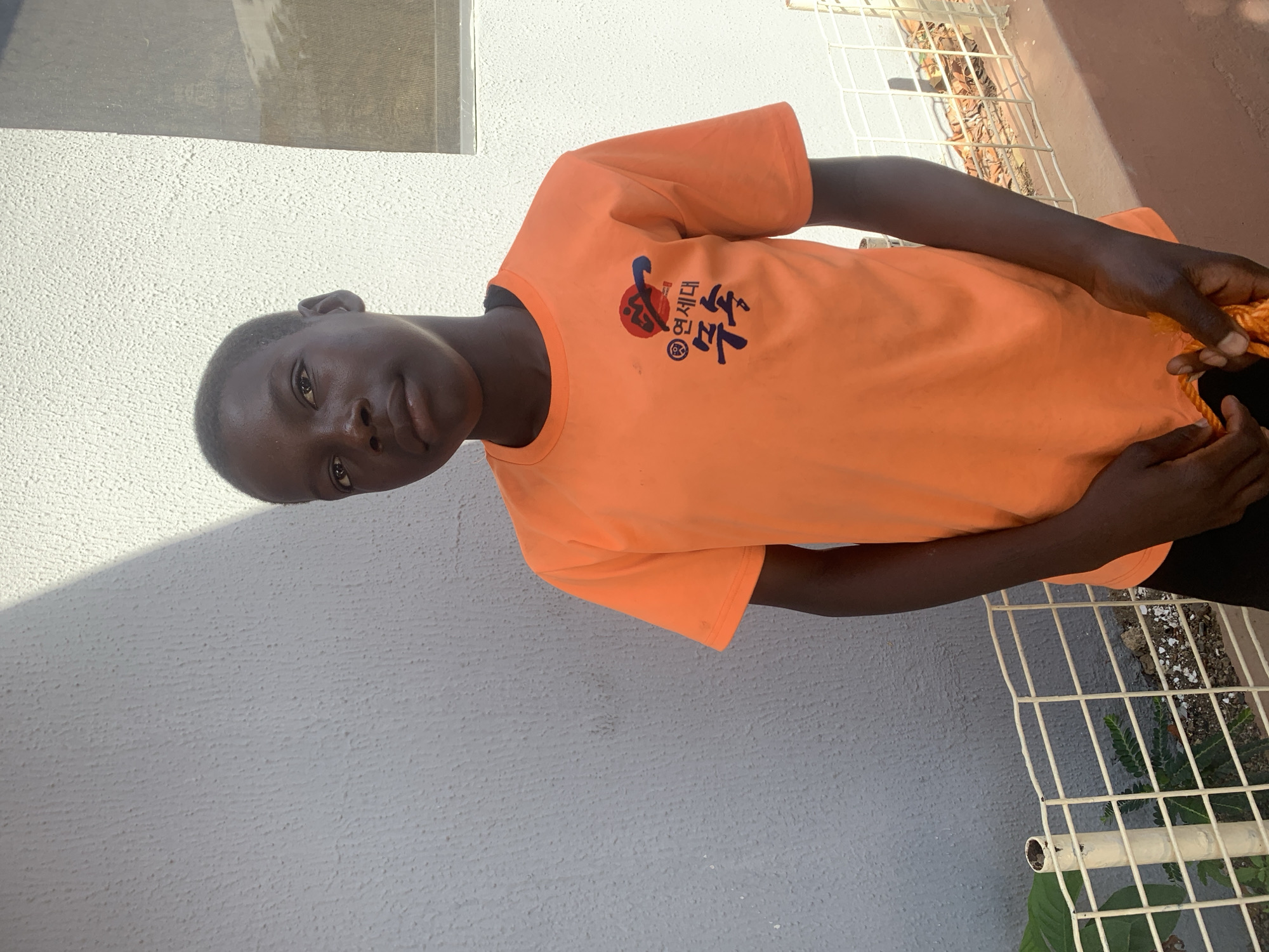 A young person stands in front of a building with a white, textured wall and a partially visible window, next to a white metal fence with vegetation growing beneath it. The person is wearing a bright orange t-shirt with a logo printed on the front in Korean script. The location is plausibly within Jos, Nigeria, given the style of the building and environmental details. The individual is holding an orange object, possibly rope, with both hands. The environment is partly shadowed, suggesting daylight.