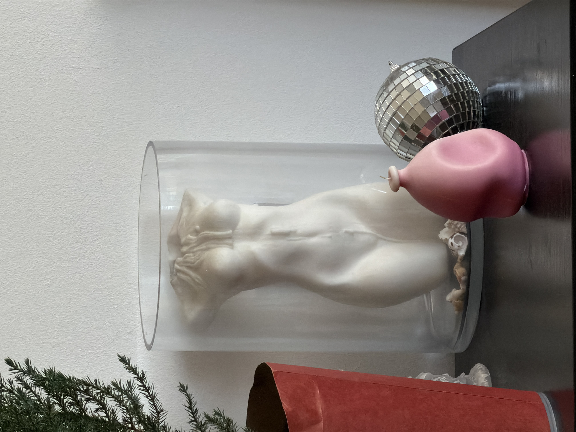 The image shows a white torso sculpture encased in a clear cylindrical tube, lying horizontally on a dark surface. Beside the tube are a disco ball and a deflated pink balloon. Below, partially visible, is a red object, possibly a box. A green coniferous plant is present in the bottom left corner, while the background consists of a white wall transitioning to the dark surface.
The image does not provide sufficient information to ascertain the location in Cape Town, South Africa.