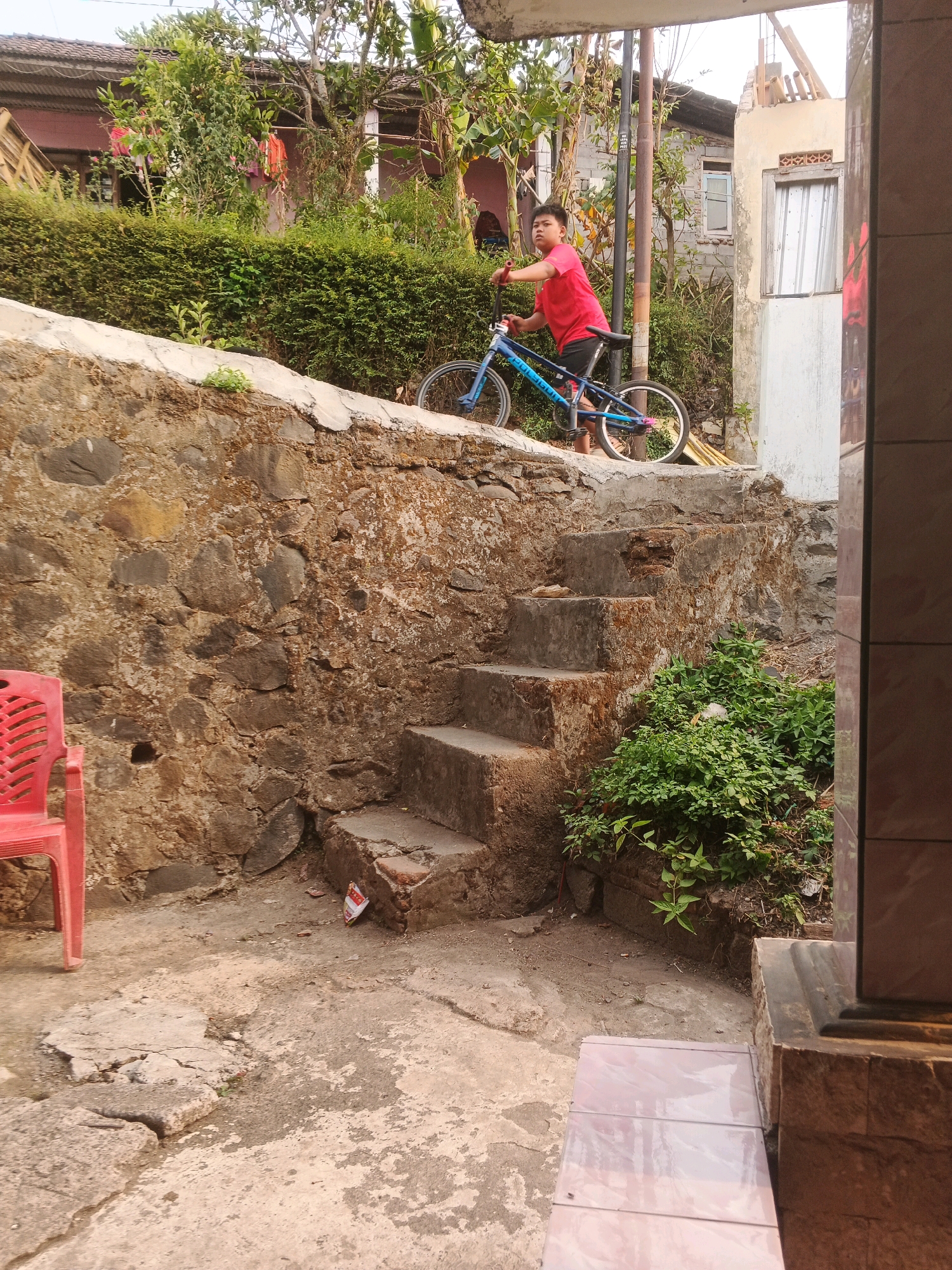 A young boy in a red shirt and black shorts rides a blue bicycle up a stone staircase in what appears to be the city of Keji, Indonesia. The boy is positioned in the middle of the frame, leaning slightly forward as he pedals. The bicycle is a BMX-style bike with a blue frame and silver handlebars. Behind the boy, a wall constructed of large, uneven stones forms the backdrop. The wall is partially covered in moss and lichen, suggesting it is old and exposed to the elements. To the right of the staircase, lush green vegetation spills over the edge of the wall. In the background, glimpses of houses with red tiled roofs and white walls can be seen through dense foliage, indicating a residential area. A red plastic chair is visible to the left of the stone wall. The ground is a mix of concrete and dirt, with some cracks and uneven surfaces. The lighting suggests it is daytime, with the sun casting shadows and highlighting textures.