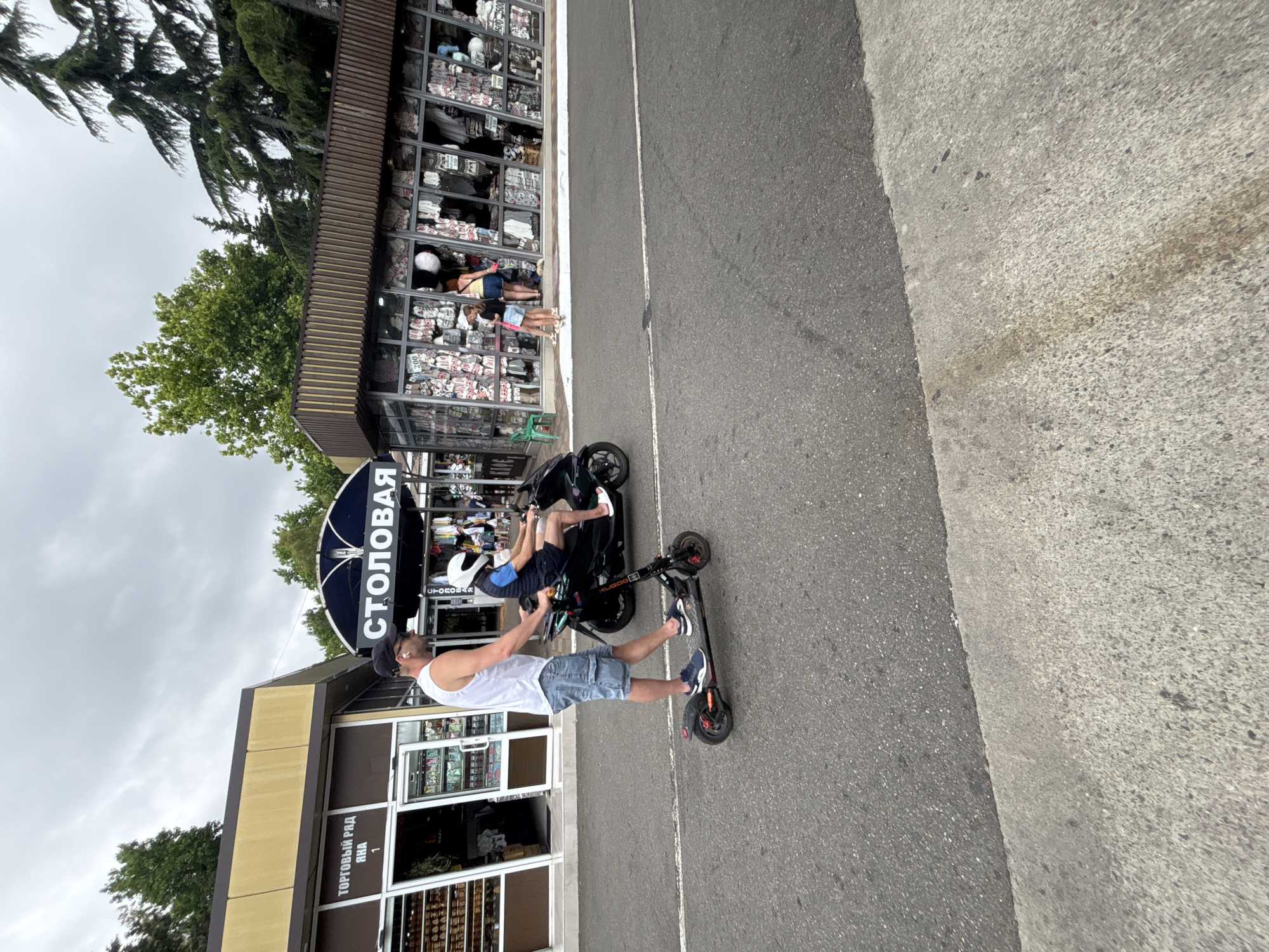 In Sochi, Russia, on a cloudy day, a man wearing a white tank top and denim shorts rides an electric scooter on an asphalt road. He is pushing another man, who is seated on a scooter and wearing a blue t-shirt and shorts with a white helmet. Both men are moving from left to right. In the background, there are several market stalls and shops. One stall, with a yellow awning, has Russian text that reads "ТОРГОВЫЙ РЯД" (Trading Row). Another stall has "СТОЛОВАЯ" (Canteen) written in large letters on its dark blue awning. The shops display various goods, including what appears to be clothing and souvenirs. Several trees, including pines and deciduous trees, are visible behind the stalls. The road is bordered by a concrete walkway on the right side.