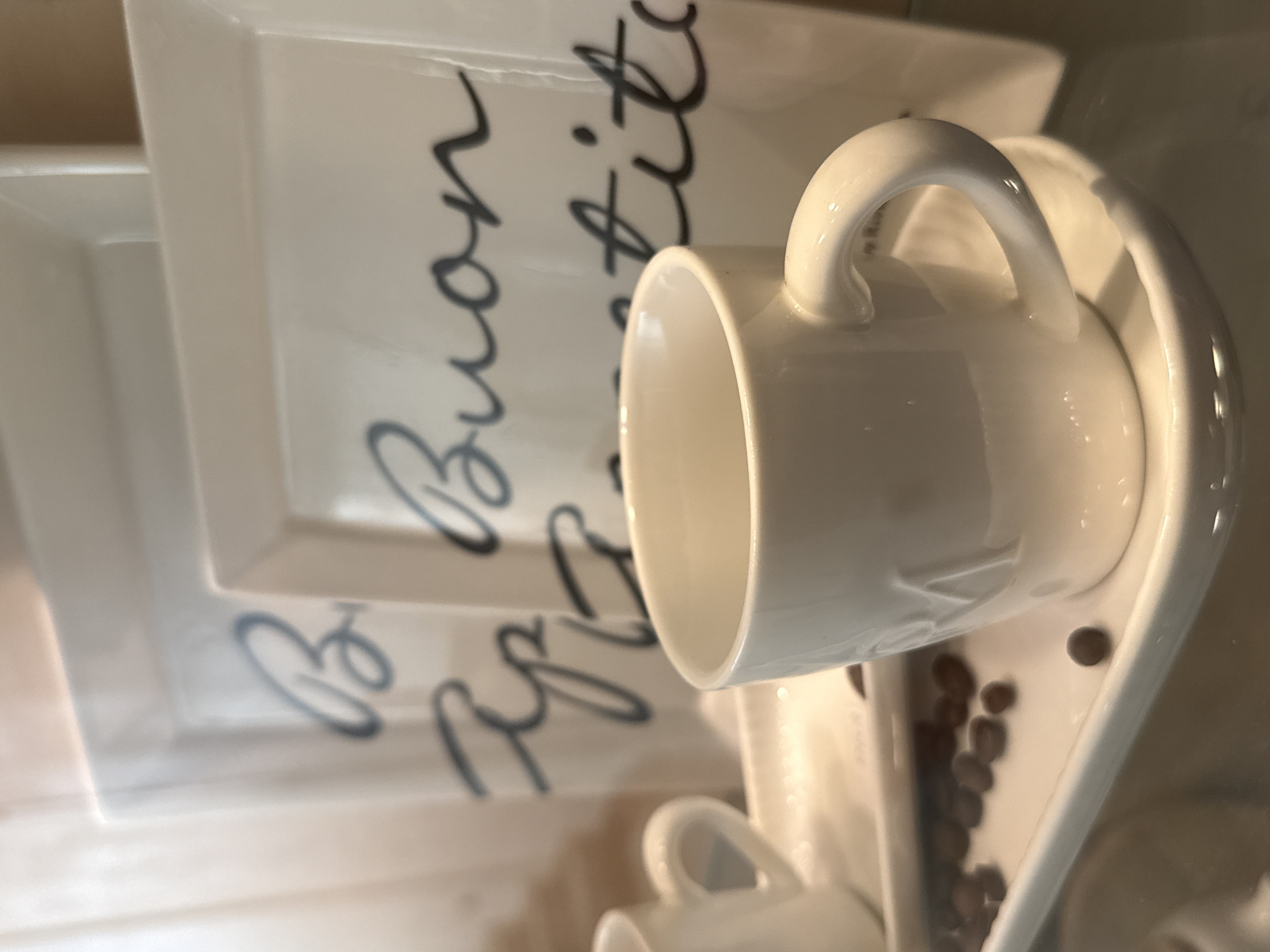 The image shows a close-up of a white ceramic coffee cup and saucer, with coffee beans scattered on the saucer. In the background, there are two white square plates with black cursive writing that reads "Buon Appetito". The scene appears to be indoors, possibly in a cafe or restaurant. The lighting is soft and warm, creating a cozy atmosphere. The image does not contain any information about the city of Bad Sachsa, Germany.