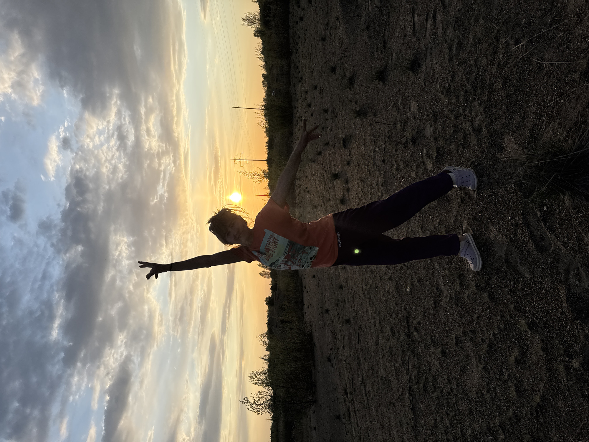 A person is striking a pose against a sunset backdrop. The individual, dressed in a tee-shirt and trousers, stands on a dirt surface and extends both arms outward, as if balancing. The location appears to be outdoors in a natural setting, possibly near Noviy, Russia. The sun is setting on the horizon.