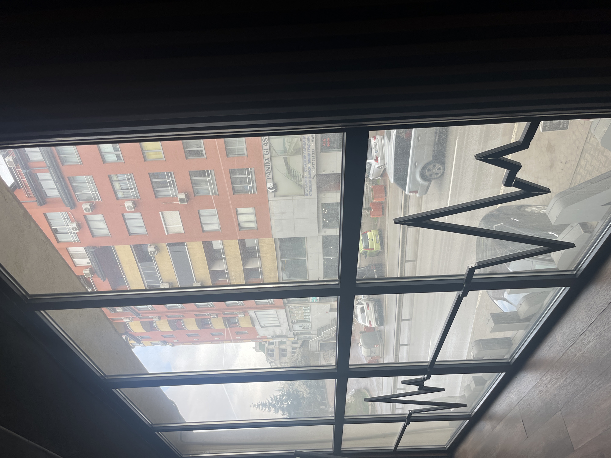 The image shows an interior view through a multi-pane window, revealing an urban scene in Sofia, Bulgaria.
The foreground is defined by the window frame, which holds multiple panes of glass. Attached to the frame are dark, zigzag-shaped metal rods, possibly part of a curtain or shade mechanism. The indoor environment appears dim, contrasting with the brighter outdoor scene visible through the window. The interior floor is visible in the bottom right corner, showcasing a dark wood finish.
The exterior view displays an urban landscape. A multi-story red and yellow building stands across the street, showing rows of windows and air conditioning units. Adjacent to it, a taller building features a business called "PANDA GLAS" with accompanying signage, indicating the company sells glass products. The street below is filled with vehicles, including cars, a truck, and other unidentifiable vehicles. Pedestrian walkways and patches of greenery are also visible. The weather appears overcast.