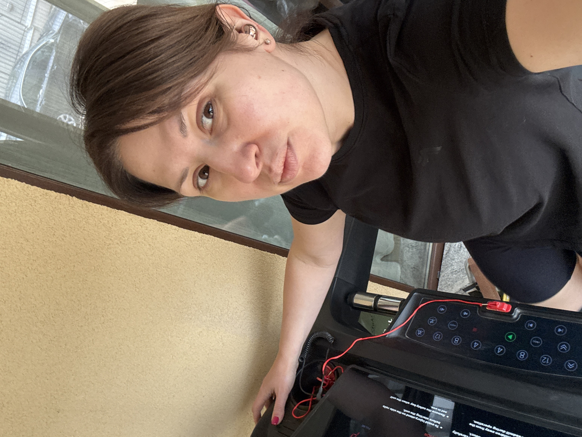 The image shows a woman with brown hair and a black t-shirt standing next to a treadmill. She is facing the camera with a slight pout. The treadmill has a display panel with buttons and text. The background includes a yellow wall, a window, and possibly an outdoor area. There is no location context within the image. I cannot provide the location in the city of Sofia, Bulgaria based on the information presented.
