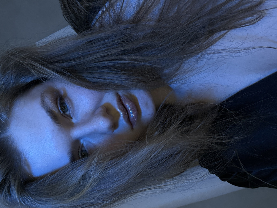 A woman lies with her head tilted. Her light-brown hair is spread around her face and upper chest. She is gazing at the viewer. The color palette consists of blues, grays, and browns. The lighting is predominantly blue, casting shadows and highlighting her features. The visible portion of her clothing is a dark color, possibly black.