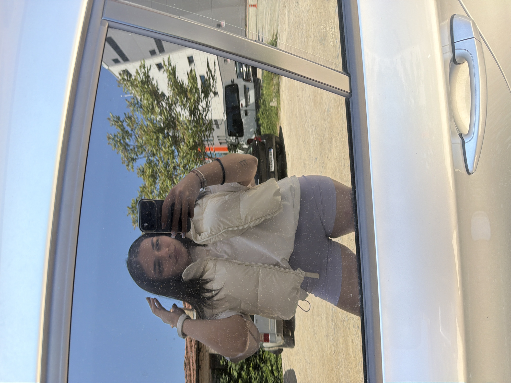 A mirror selfie captured on a smartphone shows a young woman with dark hair reflected in a vehicle window. She is wearing a white vest over a white shirt and lavender shorts. Her right arm is raised to hold the phone. The reflection in the car window shows a building, a tree, and other vehicles in the background. The license plate on one vehicle in the background reads “EB 3795 BH.” A light-colored car door is visible on the right side of the frame. Based on the license plate, the location is Gabrovo, Bulgaria.