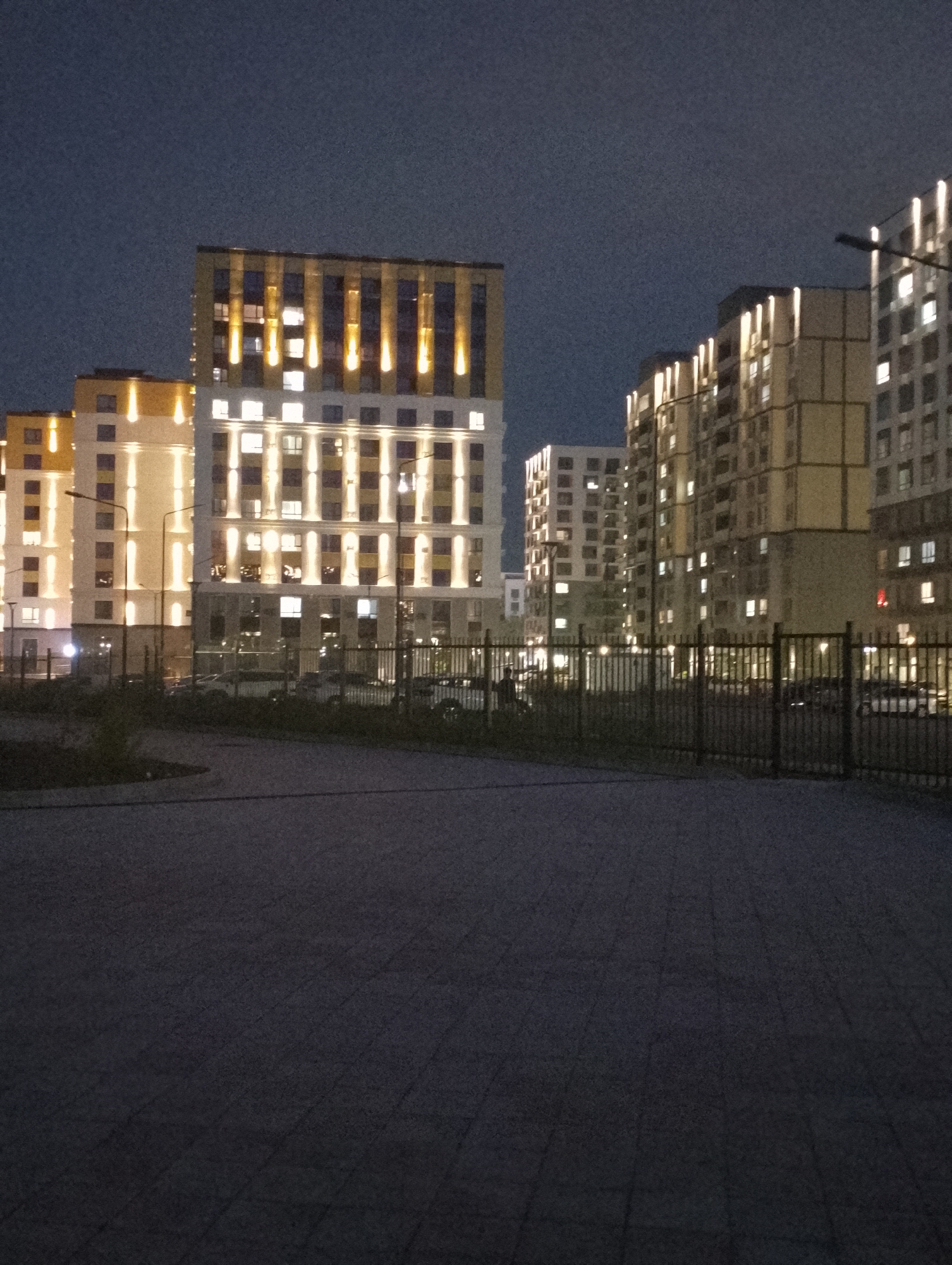 The image depicts a nighttime view of a modern residential complex in the city of Almaty, Kazakhstan. Several multi-story apartment buildings are visible, illuminated by exterior lights and interior lighting from windows. The buildings have a contemporary architectural style with clean lines and a light-colored facade. A dark metal fence runs across the foreground, separating a paved area from the buildings. In the background, vehicles are parked, and streetlights are present. The sky is dark, indicating it is nighttime. The overall scene suggests a well-lit, developed urban area.