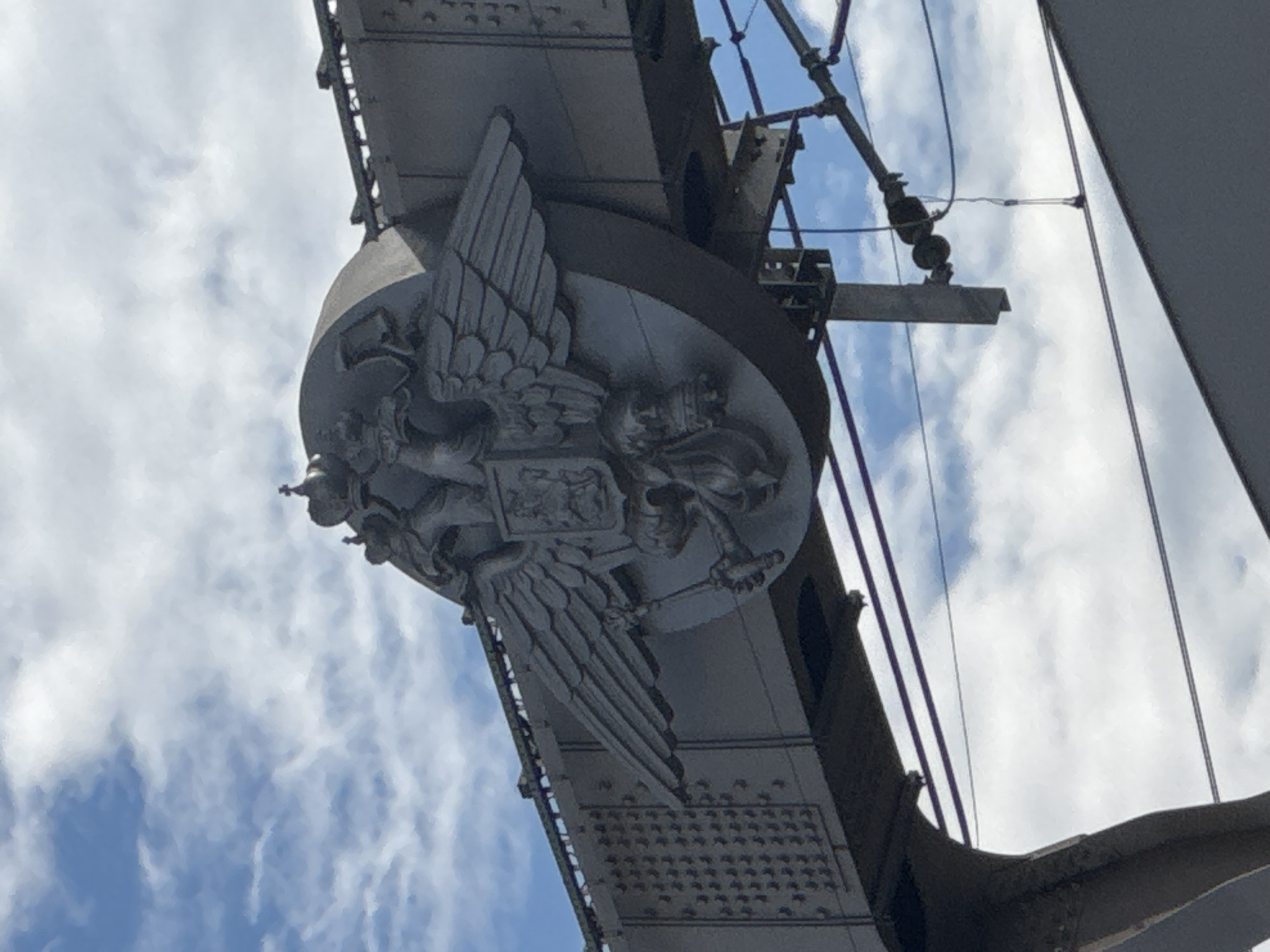 The image shows a close-up, slightly tilted, low-angle view of a large, ornate metallic emblem attached to a bridge structure. The emblem features a prominent double-headed eagle, a common symbol of Russia, with detailed wings, talons, and a shield on its chest. The eagle's heads are facing outwards, and its wings are spread wide. The entire emblem is cast in a dark, weathered metal, giving it a somber and historic appearance.
The emblem is affixed to a massive, industrial-looking bridge, characterized by its heavy metal beams, rivets, and supporting structures. The bridge's framework is visible in the background, suggesting a substantial piece of engineering. Several wires and cables are also visible, possibly part of the bridge's electrical or support systems.
The sky behind the bridge is a mixture of bright blue and fluffy white clouds, indicating daylight, likely midday or early afternoon. The lighting suggests a sunny day, though the clouds create some diffused light.
While no people are visible, the scale of the emblem and the bridge implies a public space, likely a significant thoroughfare or landmark in Moscow, Russia. The overall impression is one of historical weight and enduring strength. No text is discernible in the image.
