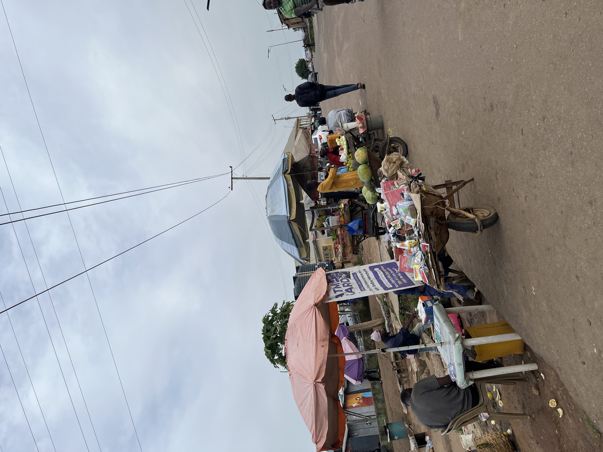 The image depicts a bustling street scene in Gura Top, Nigeria. The main subjects are several people engaged in commerce and pedestrian activity. A fruit vendor has set up a colorful display on a cart filled with watermelons, pineapples, and other produce. The cart is adorned with various goods for sale, including bottled drinks and other packaged items. Two large umbrellas, one pink and orange and another blue, provide shade for the vendors and their wares. In the background, other individuals are walking along the paved road, and some vehicles are visible, suggesting a lively marketplace.
The scene appears to be during the daytime, with overcast skies indicating potentially cloudy weather. The setting is clearly a street, characterized by the asphalt road and overhead power lines. One of the vendors is interacting with customers, while others are attending to their stalls. The overall atmosphere seems active and commercial.
Several pieces of text are visible in the image, primarily on signs and product packaging. One prominent sign advertises "ATM CARD" with details about "Maintenance Fees," "Online Merchants," and "Free Bank Fran." Other text on various items indicates product names and promotions. The presence of these signs further emphasizes the commercial nature of the depicted scene.