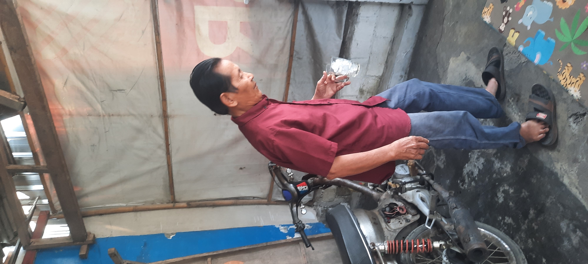 In Selopuro, Indonesia, an elderly Asian man wearing a maroon button-up shirt and blue pants is standing next to a motorcycle. He holds a glass of ice in his right hand and appears to be smoking a cigarette, with smoke rising near his face. The man is looking down and to his right. The motorcycle is partially disassembled, with its engine exposed. The scene takes place in an open-air, rustic structure with a corrugated metal roof supported by wooden beams. The walls are covered with a white tarp. In the foreground, a colorful children's play mat with animal motifs is visible on the concrete floor. The floor is dark and appears to be stained.