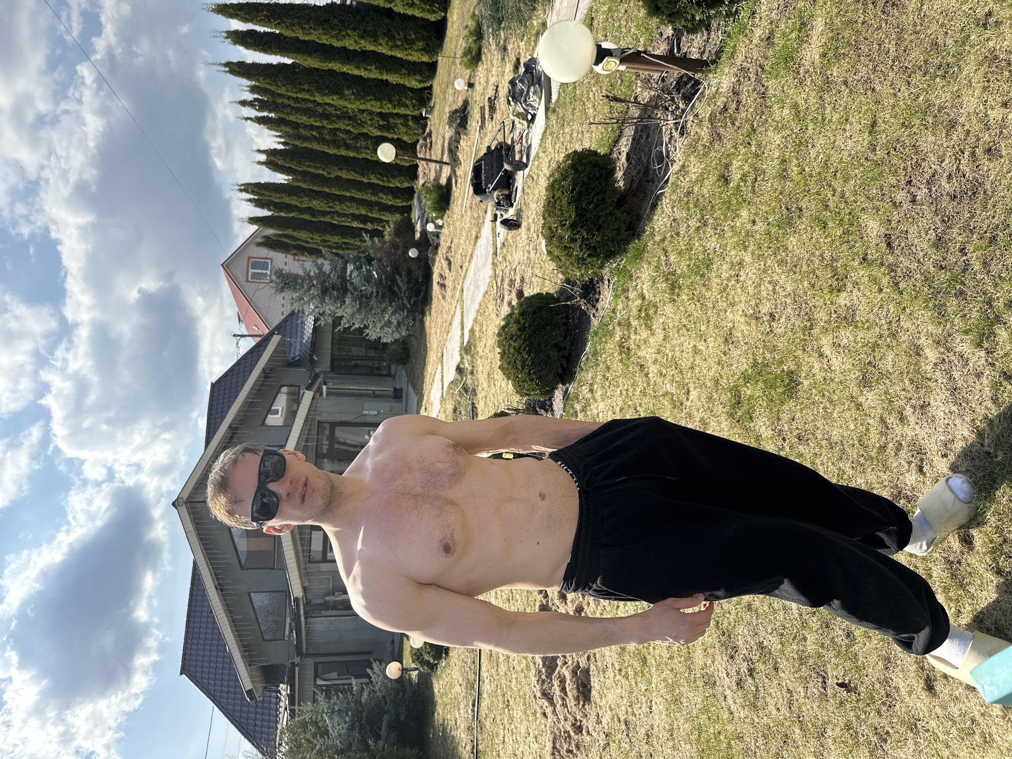 A shirtless man wearing black pants, socks, and slippers, and sunglasses, stands in a yard in Kursk, Russia. A house and trees are in the background. The yard has grass. The sky is cloudy.