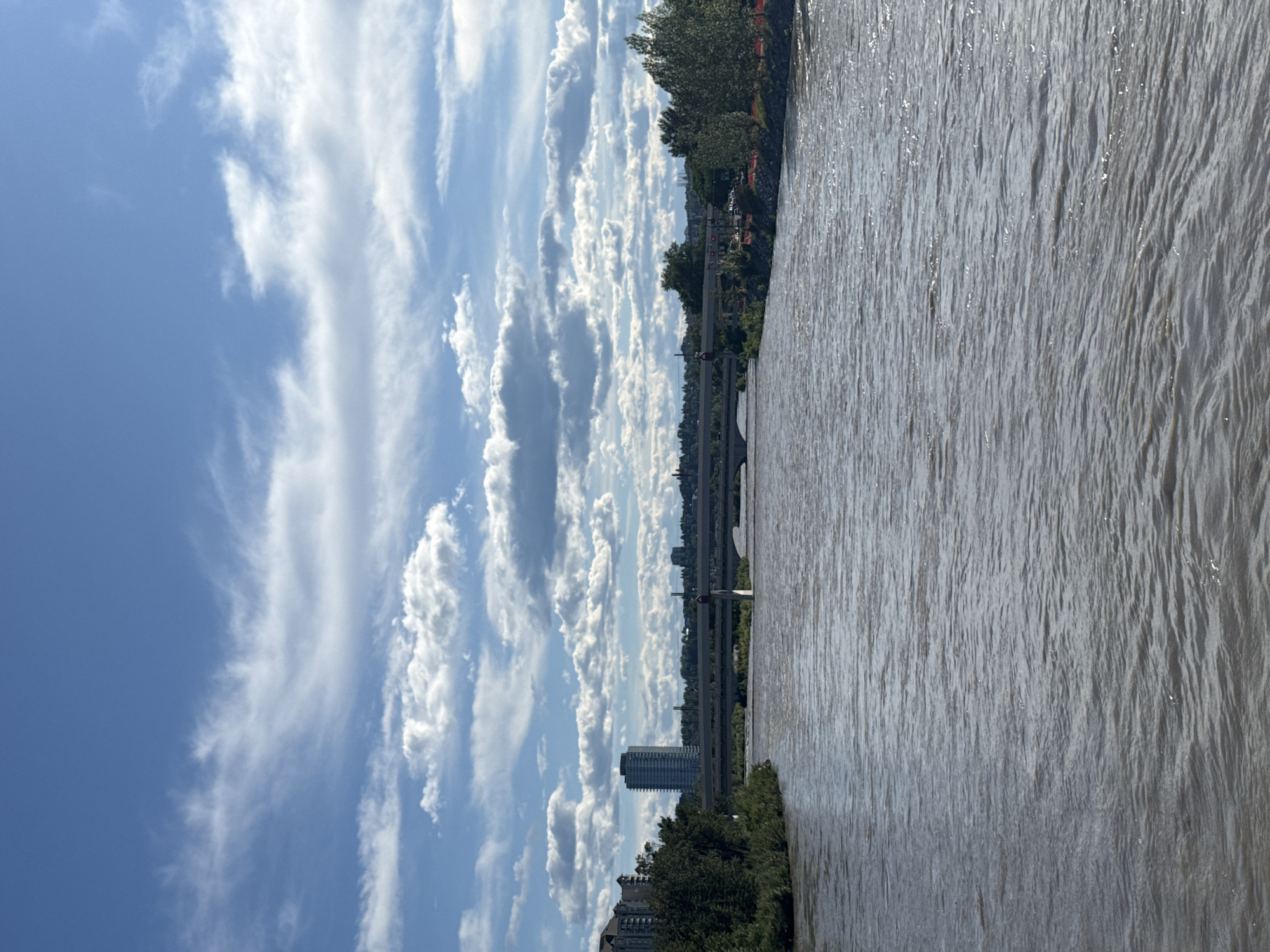 This is a wide-angle, eye-level, outdoor photograph of the Bow River in Calgary, Canada, taken on a sunny day with scattered clouds. The river occupies the majority of the foreground, its surface disturbed by ripples and reflecting sunlight. In the middle ground, a multi-lane highway bridge spans the river. Beyond the bridge, a cityscape with various buildings, including a prominent tall, slender, dark-colored building and other lower-rise structures, is visible in the distance. The banks of the river are lined with trees and greenery. The sky is a clear blue, populated by numerous white, fluffy cumulus clouds, some of which appear thin and wispy. The overall impression is a scenic view of an urban river with a bridge and city skyline under a dynamic sky.