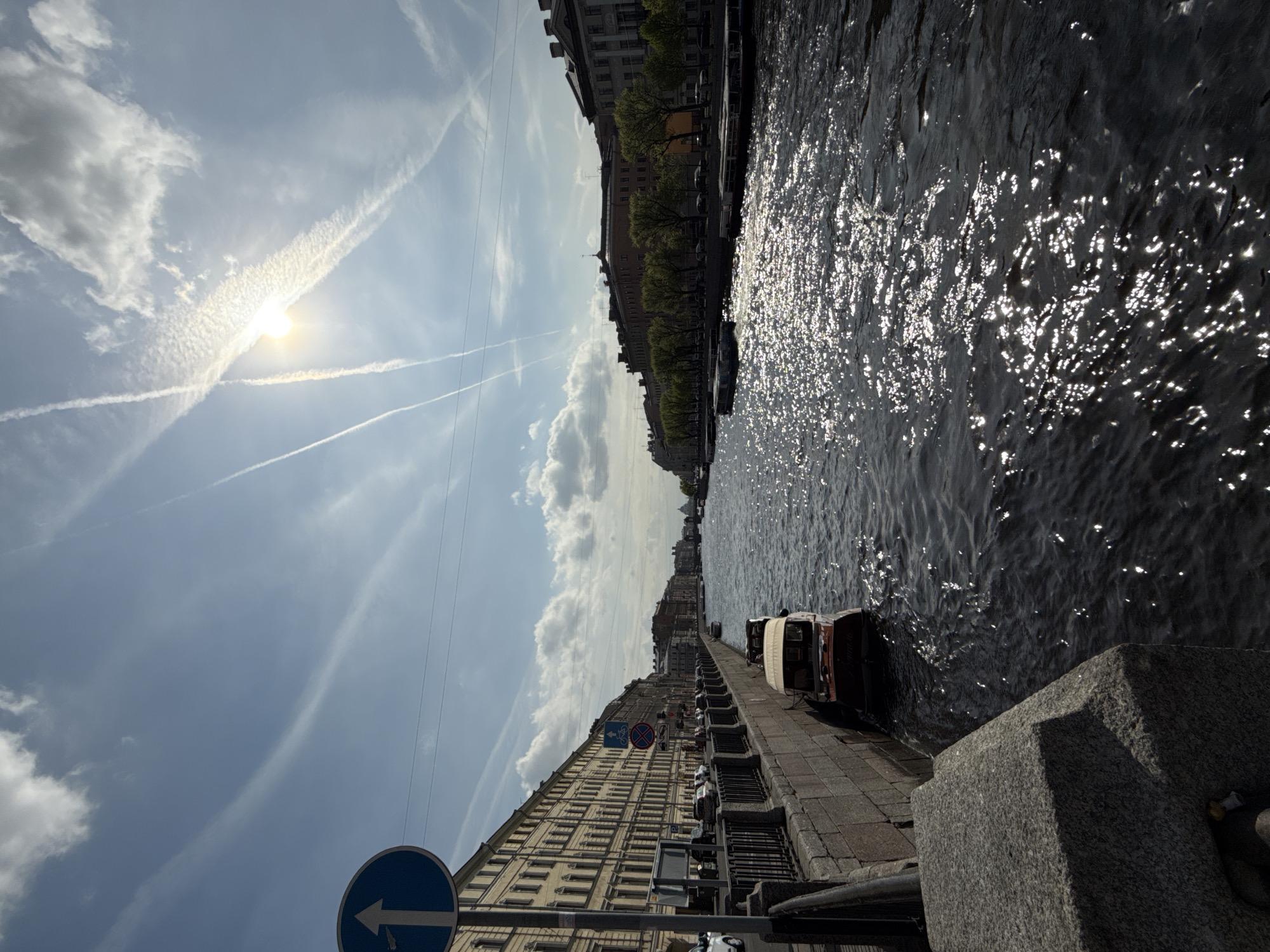 The image shows a waterway, likely a canal or river, in Saint Petersburg, Russia. Buildings line the waterway, their architecture characteristic of the city. The sky is partly cloudy with contrails visible. A boat is docked along the embankment. A road sign with a directional arrow is visible on the lower left of the image. Sunlight reflects off the water's surface.