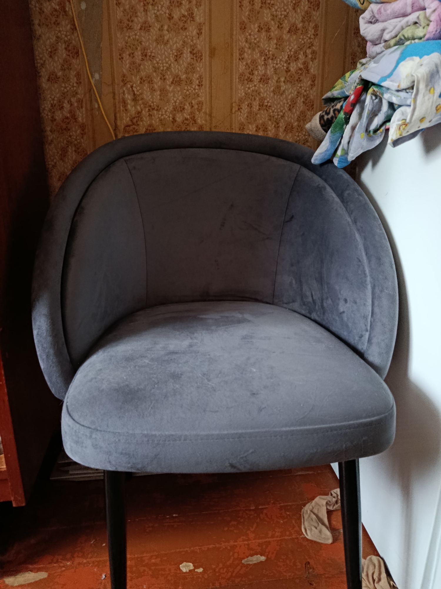 The image depicts an interior scene with a dark gray upholstered chair as the central subject. The chair has a rounded back and a seat with visible stitching. It is supported by thin black legs. To the left of the chair, a dark wooden cabinet is partially visible. The wall behind the chair is covered in patterned wallpaper in shades of beige and brown, with a yellow wire running vertically along it. To the right of the chair, a stack of folded colorful fabric items, possibly blankets or bedding, is piled on a white surface, which could be a piece of furniture or an appliance. The floor appears to be made of wooden planks, painted a reddish-brown, with some scuff marks and peeling paint. Two beige fabric items, resembling stockings or tights, are lying on the floor beneath the chair. The lighting suggests an indoor environment, possibly a domestic setting. The image does not provide any specific information about the city of Vladimir, Russia, or any observable actions or interactions.