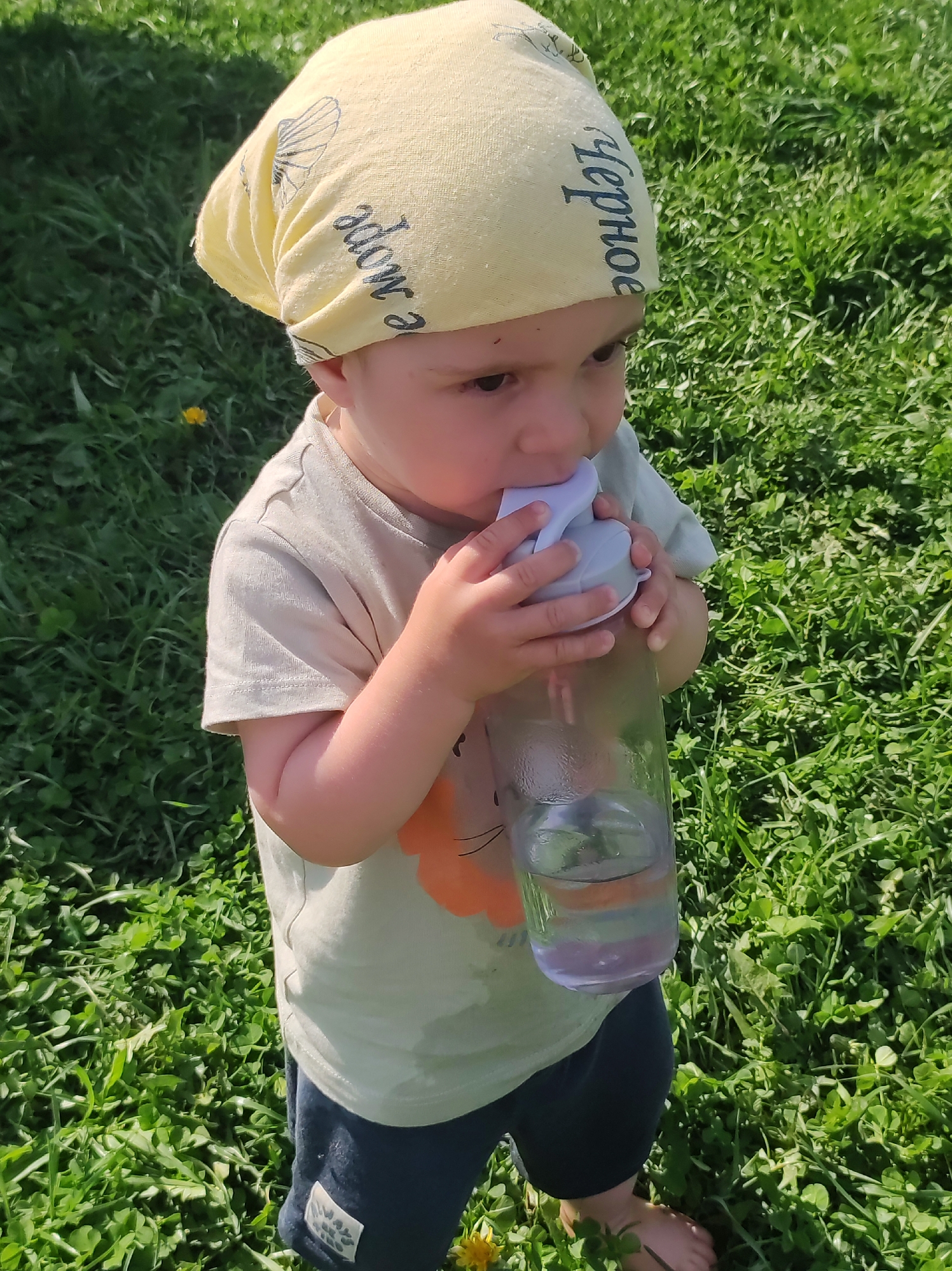 A child wearing a light yellow headscarf with black text that appears to say "Черное море", a light gray t-shirt with an orange lion design, and dark blue shorts is drinking from a clear water bottle with a light gray lid and spout. The child is standing barefoot in a field of green grass and clovers. Based on the text on the headscarf and the location provided, the scene likely takes place near the Black Sea in Фоки, Russia. The sunlight suggests it is daytime.