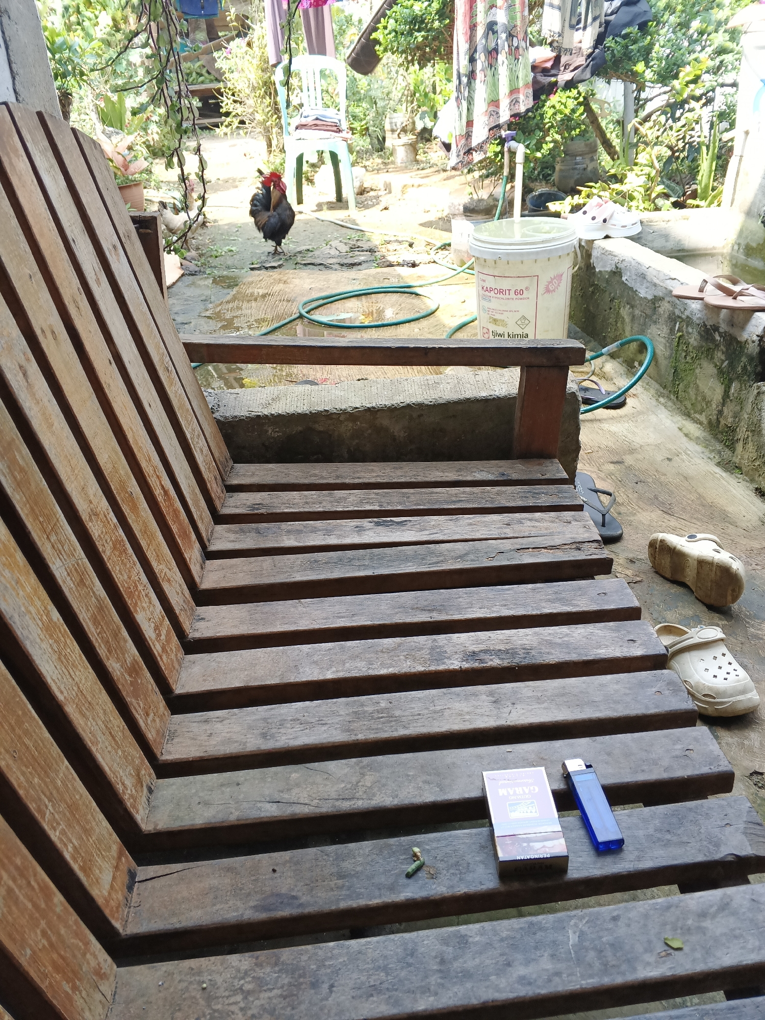 A rooster stands on a wet, uneven concrete ground surrounded by greenery. In the foreground, a wooden bench with slatted seating faces the viewer. On the bench, a pack of "GARAM" cigarettes and a blue lighter are placed next to each other. In the background, a white bucket with "KAPORIT 60" written on it sits near a faucet and a coiled green hose. A pair of white clogs and brown flip-flops are visible near a concrete structure, suggesting an outdoor, possibly rural setting. The overall scene suggests a humid environment, likely in Warungkiara, Indonesia, given the presence of tropical vegetation.