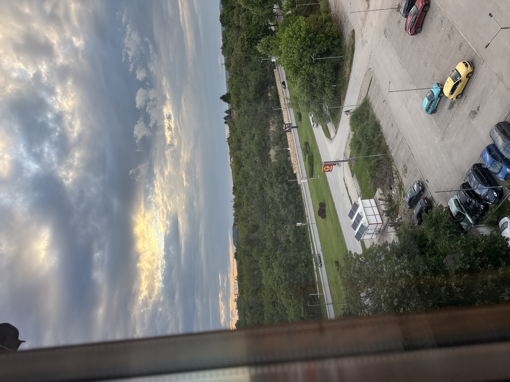 Captured from an elevated perspective, the image reveals a cityscape on a partly cloudy day. In the foreground, a window frame hints at an indoor vantage point. The view extends to a street lined with greenery and trees. A parking lot is visible, occupied by several cars of varying colors, including yellow, blue, red, and black. The environment includes architectural features like street lamps and possibly solar panels. There are no visible individuals, but the presence of cars and street lamps suggests a residential or commercial area. Given the request details, the location is likely Lovech, Bulgaria.