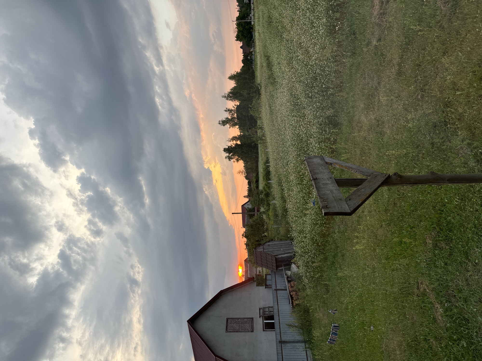 This is a vertical, eye-level photograph of a rural scene at sunset. The sky dominates the left side of the frame, filled with dramatic, heavy grey clouds streaked with the warm hues of the setting sun. A band of orange and pink light cuts across the horizon, indicating the sun is low in the sky.
The right side of the image depicts a green landscape, likely a field of grass or wildflowers, dotted with tiny white blossoms. In the foreground of this field, a weathered wooden structure, possibly part of a fence or a stand, is visible. Further back, several houses and outbuildings are nestled amongst trees. One house in the lower left is prominent, with a red roof and grey siding. The overall impression is one of a tranquil, rural evening. The time of day is clearly sunset, and the weather appears to be overcast with clearings that allow the sunset colors to show through. The setting is rural Ukraine, possibly Chemerne. There is no visible text in the image. The scene evokes a sense of peace and rustic beauty.