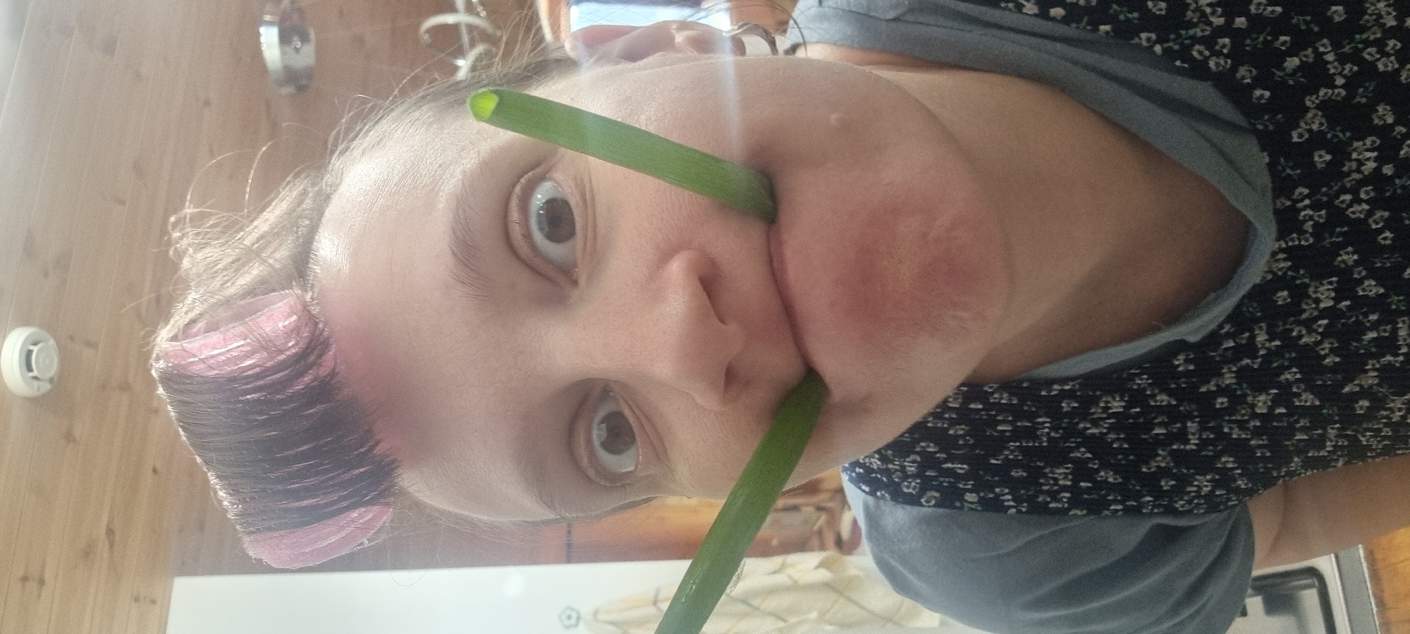 A woman is depicted with her face towards the camera, and two green onions are held horizontally across her mouth. The woman has a pink hair roller on the left side of her hair. The location is not identifiable as Dunnae-myeon, South Korea. The background includes a wooden wall, a smoke detector, and kitchen elements.