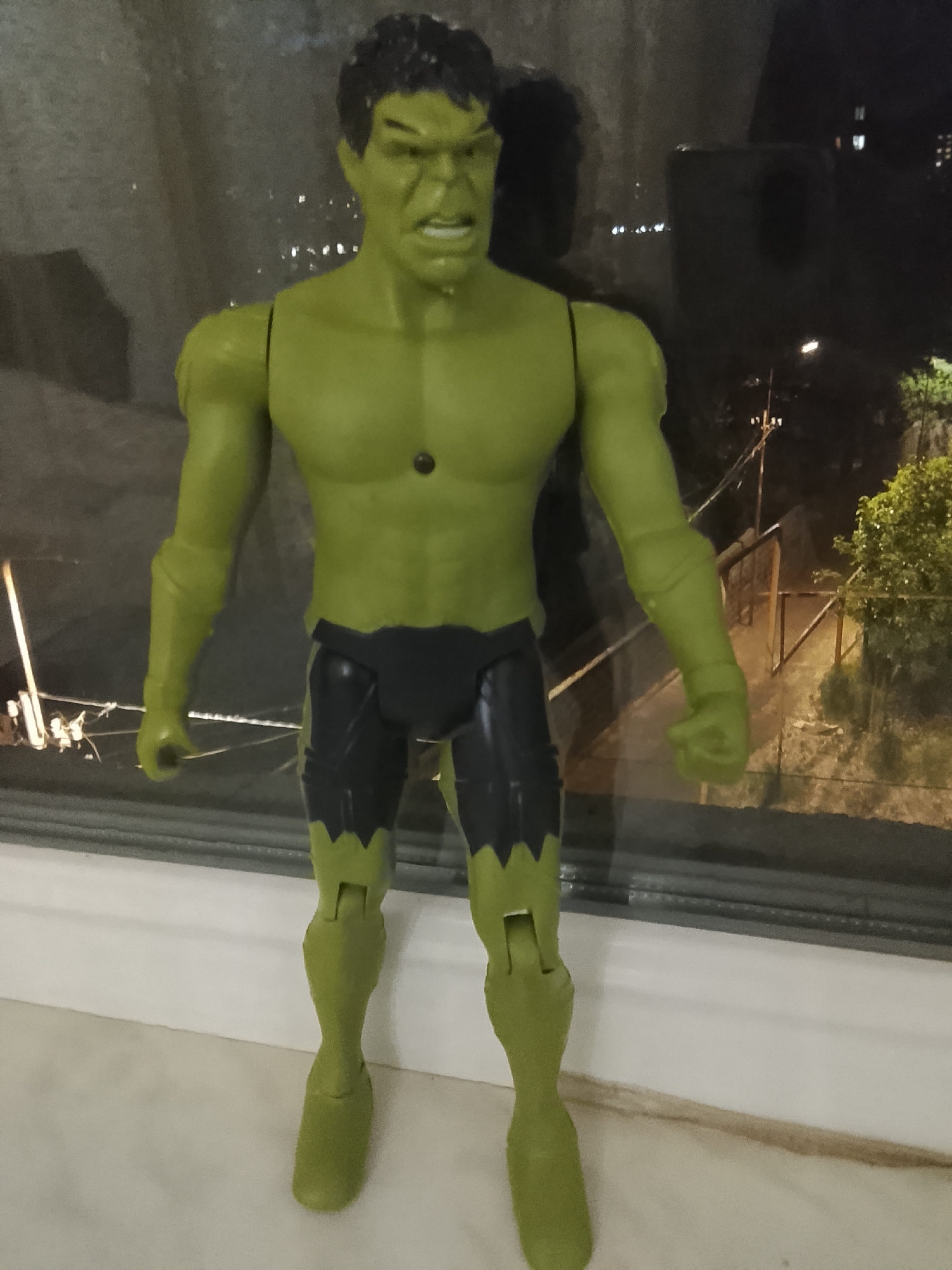 A large, green Hulk action figure stands upright on a white window sill indoors. The figure is articulated, with visible joints at the shoulders, elbows, hips, and knees. It has dark hair, a green body, and black shorts with torn, jagged green edges, typical of the character. The face of the action figure displays an angry or roaring expression, with an open mouth and furrowed brows. A small black circular detail is present on its chest. The left hand is open, while the right hand is clenched into a fist.
Behind the action figure, through a window, a nighttime scene is visible. The external environment is dark, illuminated by artificial lights from streetlights and distant buildings. There appears to be some foliage or trees, and a structure resembling a bridge or elevated walkway with railings and utility poles can be seen in the background. The window pane reflects some internal light and appears somewhat smudged or wet, creating minor distortions in the outdoor view. No visible text is present in the image.