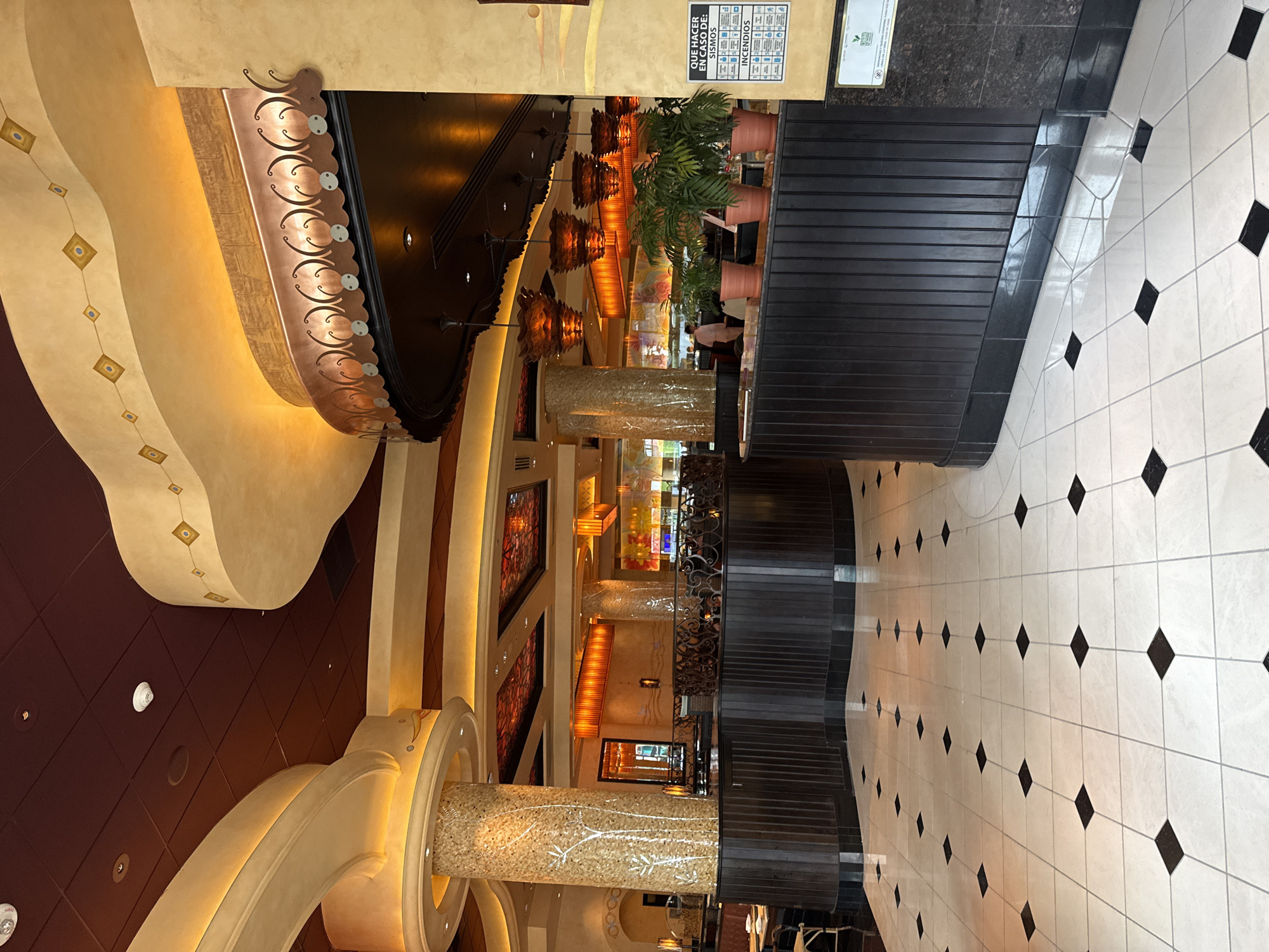 The image depicts the interior of The Cheesecake Factory restaurant, located in Mexico City, Mexico. The perspective is slightly tilted. The interior is characterized by opulent decor including tall columns, patterned ceilings, warm lighting, and decorative flourishes. The flooring is tiled in a white and black diamond pattern. A dark counter area with plants sits to the right. Several people are present, though not prominently featured. The architectural style is ornate and the overall ambiance appears upscale. A sign with instructions for emergencies is seen on a wall above the counter.