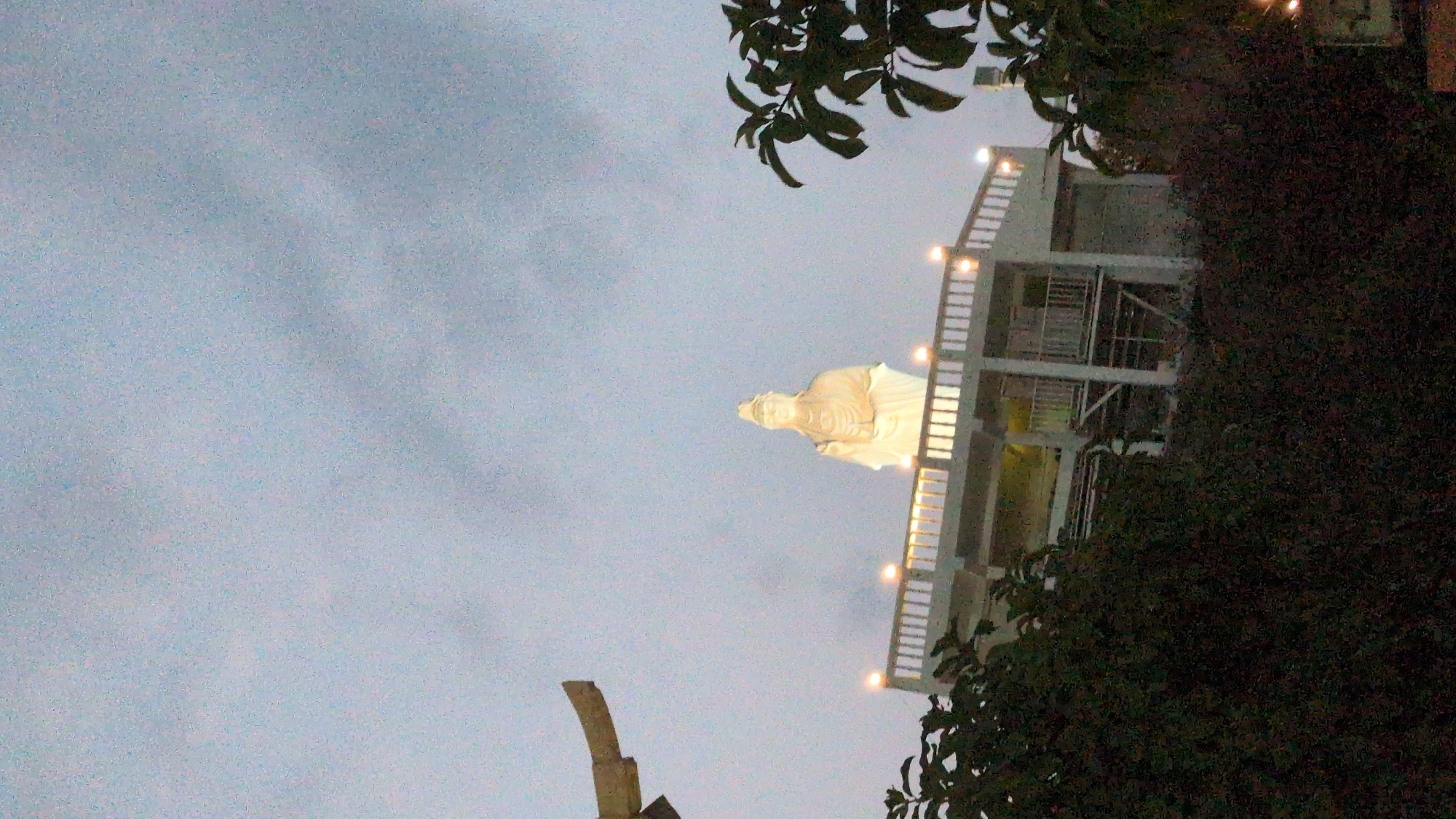 The image shows a statue on a multi-tiered structure in Khánh Hải, Vietnam. The statue, illuminated in white, is positioned on the structure's upper level. The structure itself has multiple levels with visible railings and lighting fixtures. Foliage partially obscures the right side of the structure and frames the top of the image. The sky is overcast, providing a muted backdrop. A stone structure is visible in the bottom left corner. The statue is possibly a religious figure.