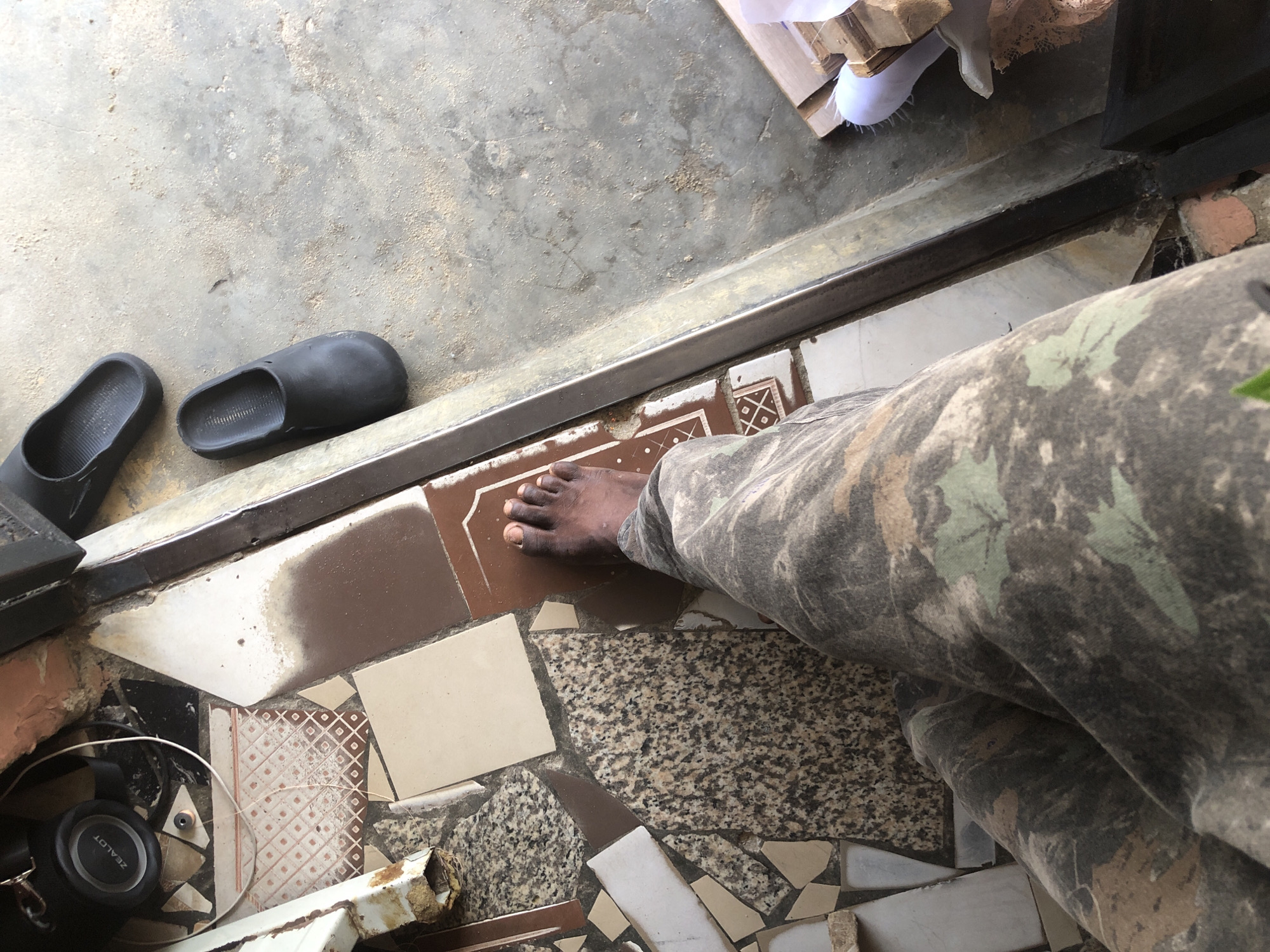 The image is a first-person perspective shot, likely taken in Ado, Nigeria, showing a person's bare left foot and lower leg clad in camouflage pants standing on a tiled surface adjacent to a concrete floor. The tiles are a mix of colors and patterns, including speckled granite, white, and brown, some of which appear broken or uneven. A metal threshold separates the tiled area from the concrete floor. Two black slip-on shoes are visible on the concrete floor, to the right and slightly above the person's foot. In the background, on the left side, a black headphone with the brand "ZEALOT" is partially visible, along with a white wire and some brickwork. The lighting suggests it is daytime.