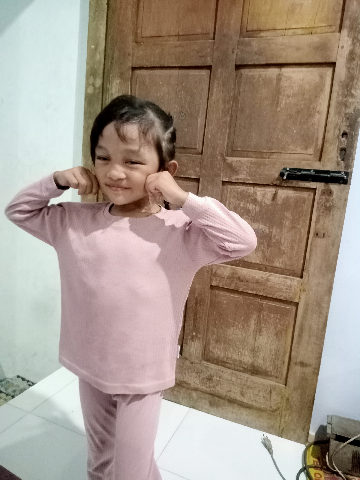 A young girl with dark hair pulled back is standing in front of a weathered wooden door. She is wearing a light pink ribbed long-sleeved shirt and matching pants. Her hands are on her cheeks, and she is smiling slightly at the camera. The background consists of a light blue wall to the left and the wooden door to the right. The floor is tiled with white squares. A black bolt is visible on the door. In the lower right corner, an electrical plug and cord are visible on the floor, along with other electronic equipment. The scene appears to be indoors, likely in a residential setting. The specific location in the city of Simo, Indonesia is not determinable from the image.