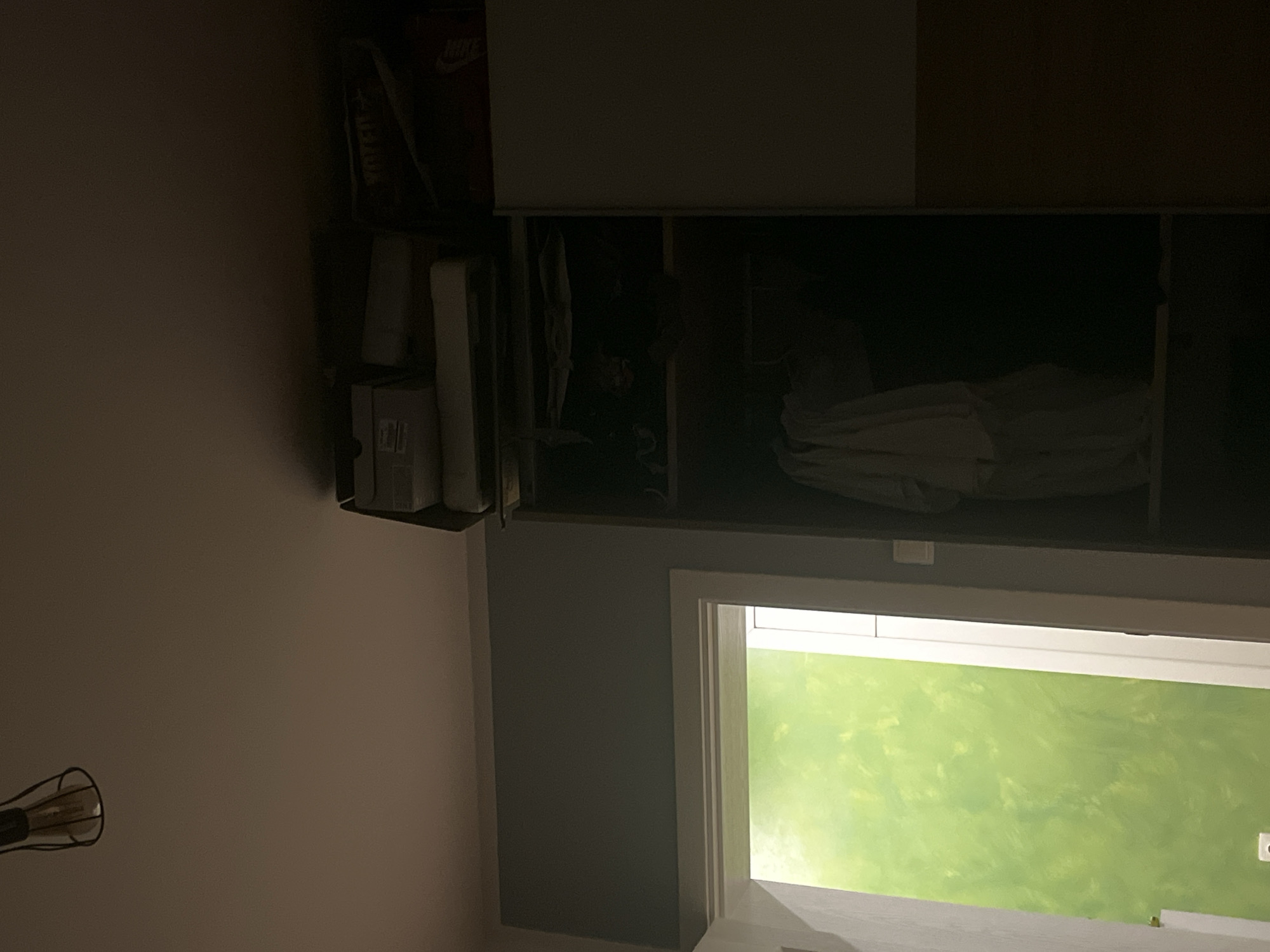 A dimly lit interior shot shows a built-in cabinet or wardrobe against a light pink or beige wall in Pokrovnik, Bulgaria. The cabinet has sections with sliding doors; the topmost section is partially visible, revealing storage above. An open cabinet section reveals assorted items, including what appears to be a Nike shoebox and various other boxes or containers. Below the cabinet, a wall with light green marbleized effect is observed. A window with white frame is located in front of the green wall. A lamp with metal cage is seen on the left side of the wall.