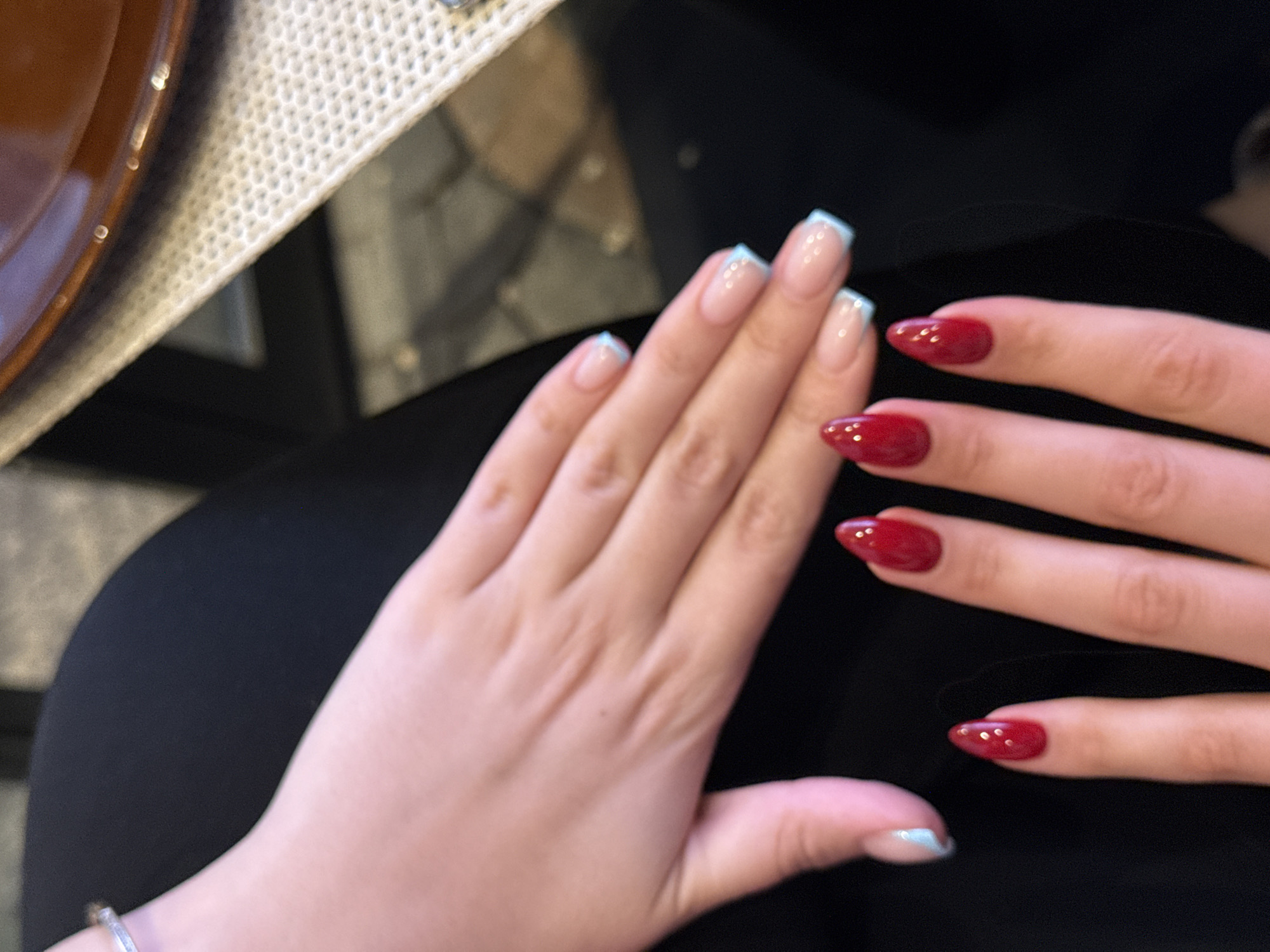 The image shows two hands with manicured nails. The left hand has a pale base with a light blue French tip. The right hand has all nails painted a dark red color. The hands are resting on a dark, likely black, material. Part of a plate and textured surface are visible in the top left corner. The location is unknown and there is no evidence to suggest it is in Yerevan, Armenia.
