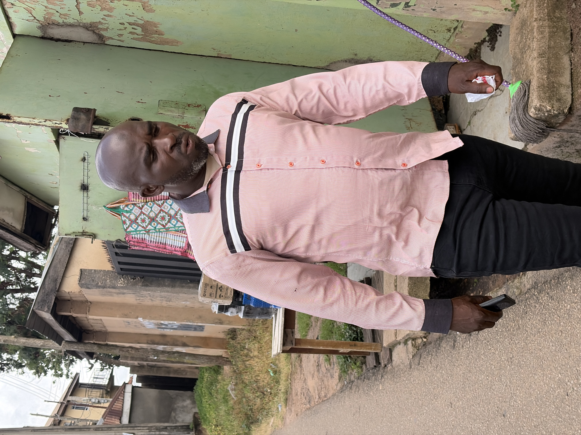 A bald Black man wearing a pink pinstripe button-down shirt with a dark collar and white stripes across the chest is walking outdoors in Jos, Nigeria. He is wearing black pants and dark cuffs on his shirt sleeves. In his right hand, he holds a white crumpled piece of paper and a mop with a purple handle and white polka dots. In his left hand, he holds a black smartphone. He is walking past a distressed light green building. Behind him, there is a tan wall with a black metal gate and some fabric bags hanging. Further in the background, there is more vegetation and buildings. The ground is a dirt path.