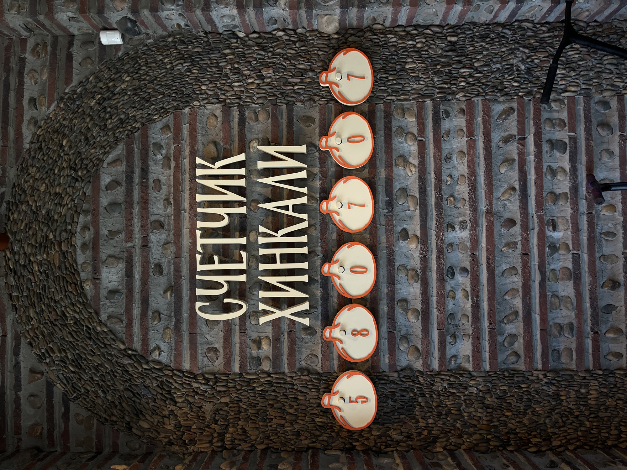 The image shows a wall with a stone archway. In the center of the wall, above a row of six circular wooden signs with numbers, are large white Cyrillic letters that spell out "СЧЕТЧИК ХИНКАЛИ" (Khinkali Counter). Each of the six circular signs displays a single orange digit. From left to right, the numbers are 5, 8, 0, 7, 0, and 7. The wall itself is constructed of horizontal brick and stone layers. The archway is filled with small, dark, rounded stones. To the right of the archway, part of a black metal object, possibly a scooter, is visible. A white cylindrical object, possibly a motion sensor or speaker, is mounted on the wall to the right of the arch. The lighting suggests an indoor environment.