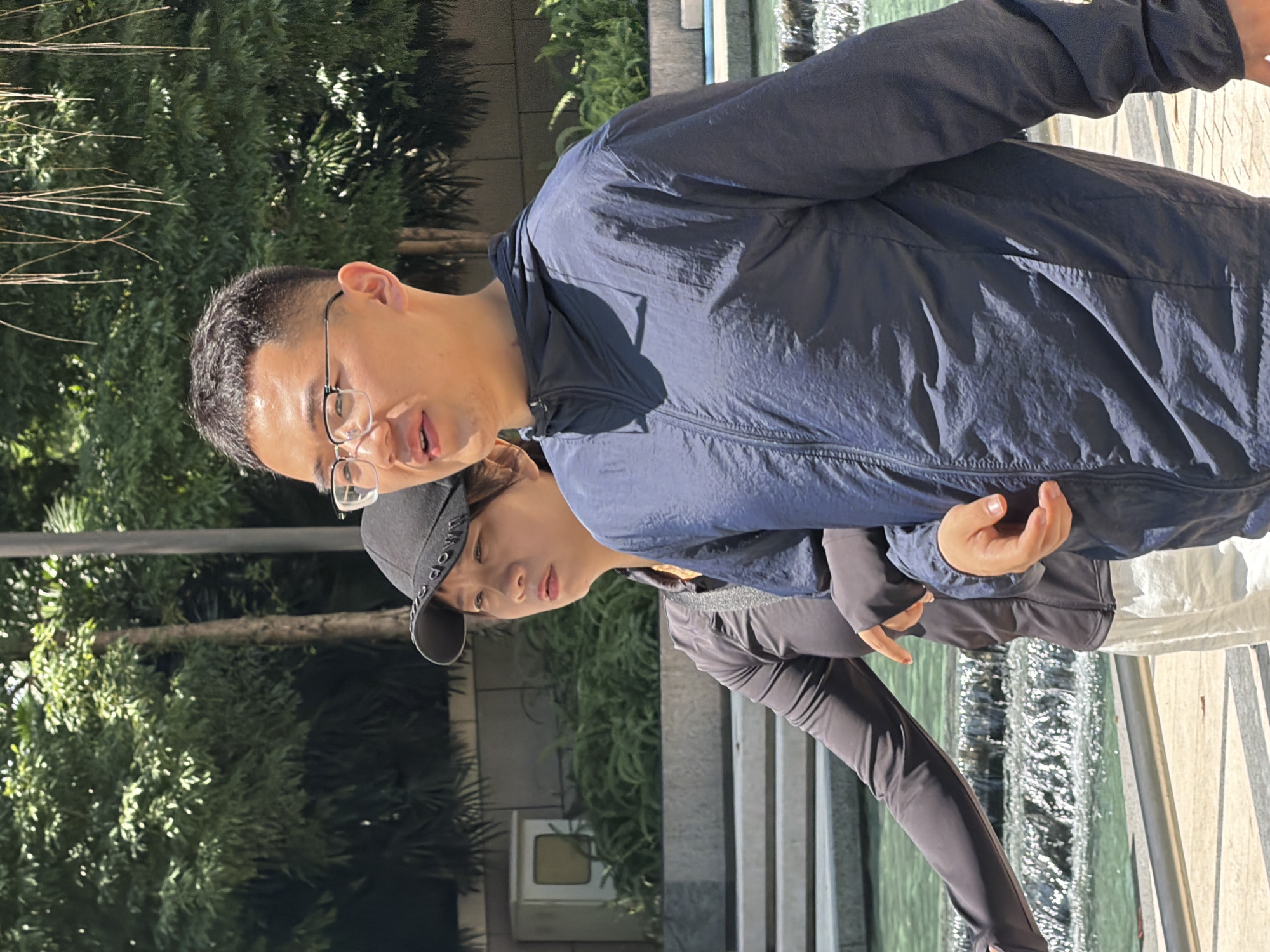 In the city of Yuzhong District, China, a man wearing glasses and a dark blue hooded jacket walks with a woman wearing a black baseball cap and a dark long-sleeved shirt. They are walking alongside a tiered water feature with lush green foliage and trees in the background. The woman has her arm linked with the man's. Sunlight filters through the leaves, casting dappled shadows.