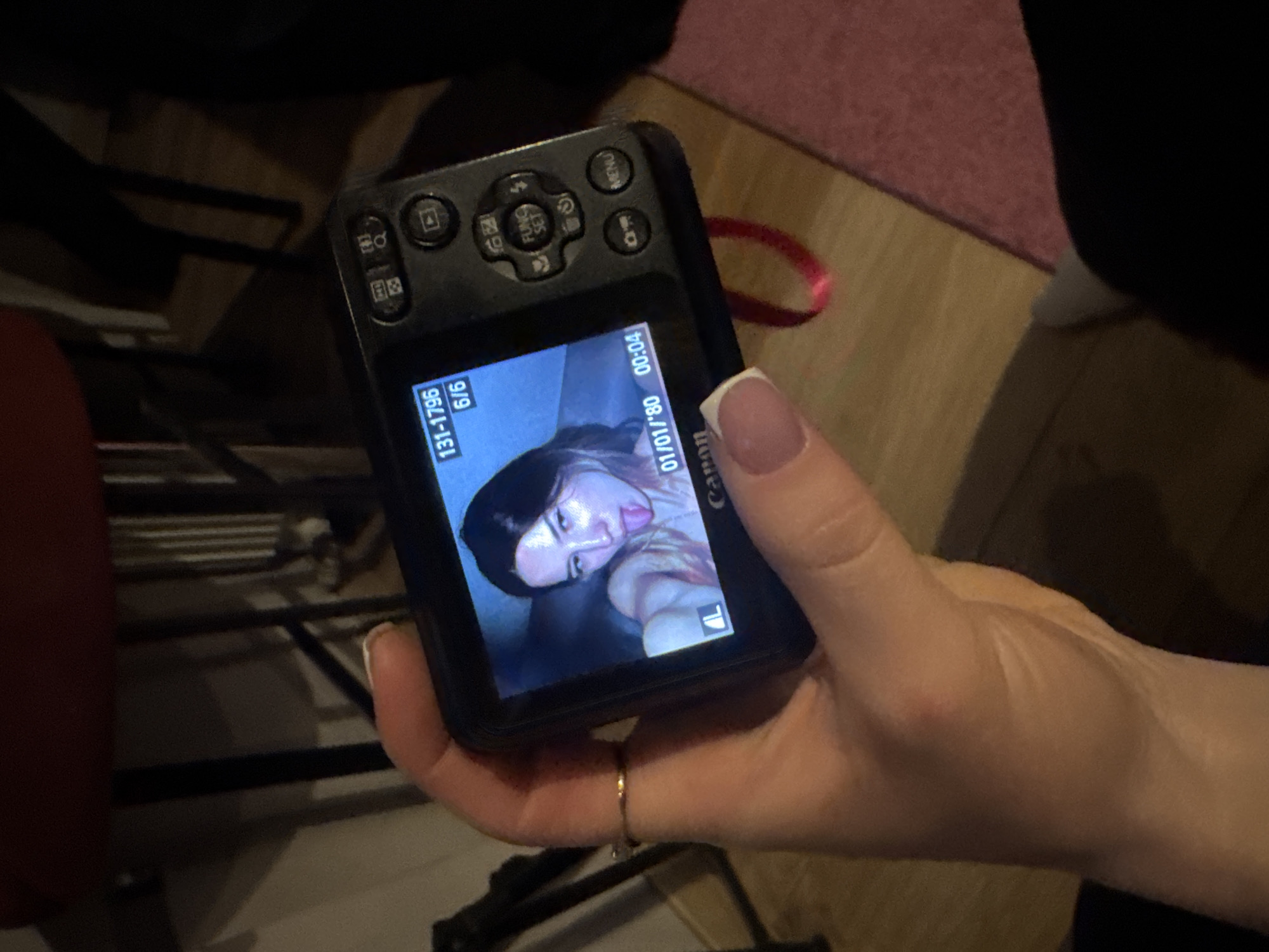 A person's hand is holding a black Canon digital camera. The camera's LCD screen displays a photo of a woman with dark hair, sticking out her tongue. The photo is dated "01/01/'80 00:04." The screen also shows "131-1796 6/6" in the top left corner. The hand has manicured nails with a white tip and a simple gold ring. The background is out of focus, but shows wooden floors, a red object, and a black metal frame. There is also a pink rug or fabric visible. The location of Khimki, Russia is not deducible from the image.
