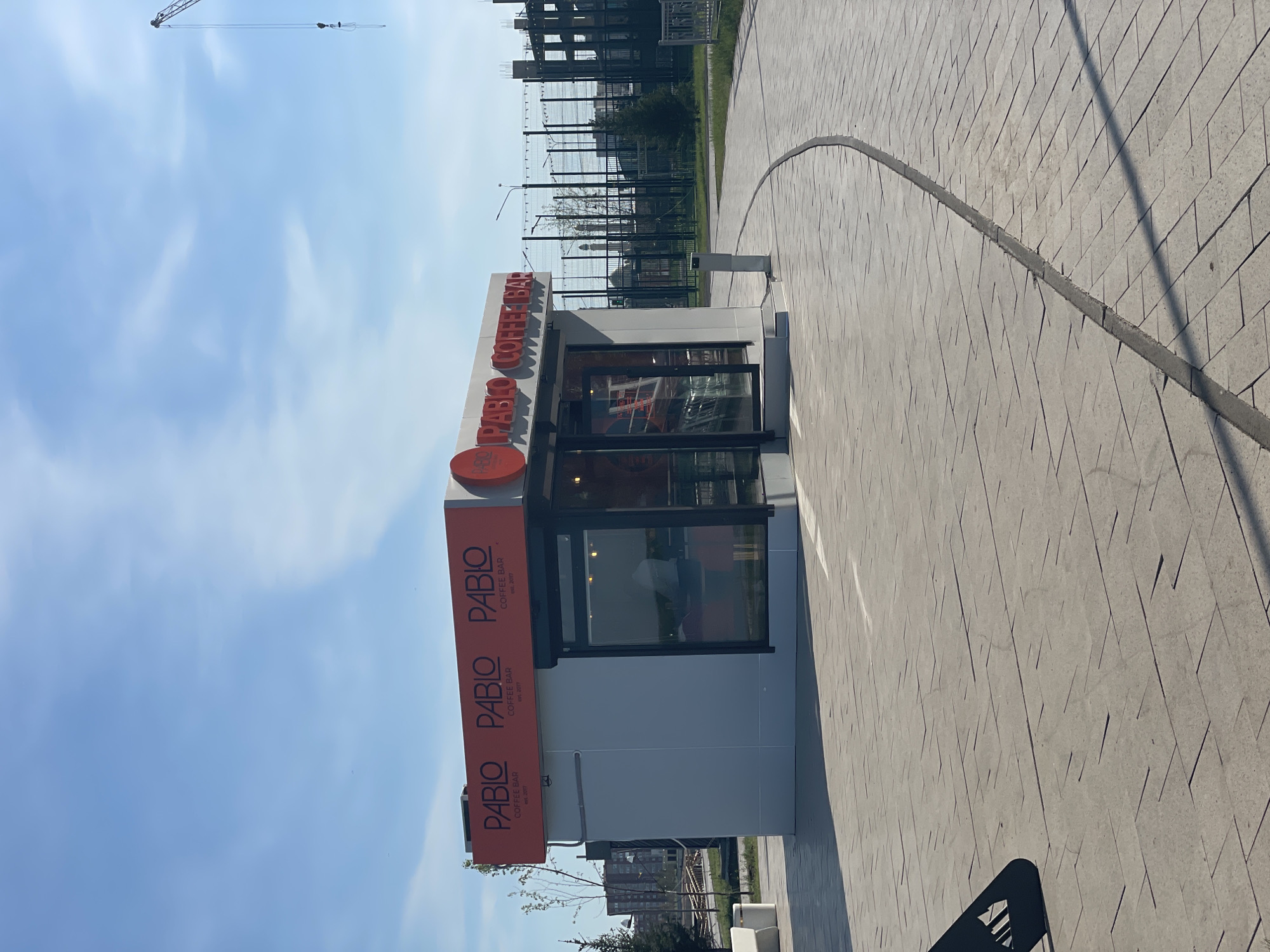 The image shows a "PABLO COFFEE BAR" kiosk located on a paved area in what appears to be Kokshetau, Kazakhstan. The kiosk is rectangular, with a white exterior and large windows. The "PABLO COFFEE BAR" sign is in orange and is prominently displayed on the top and side. The kiosk is located on a pedestrian walkway, with paving stones in a diamond-shaped pattern. In the background, there is a fence, some greenery, and buildings under construction, with a construction crane visible. The sky is blue with scattered clouds.