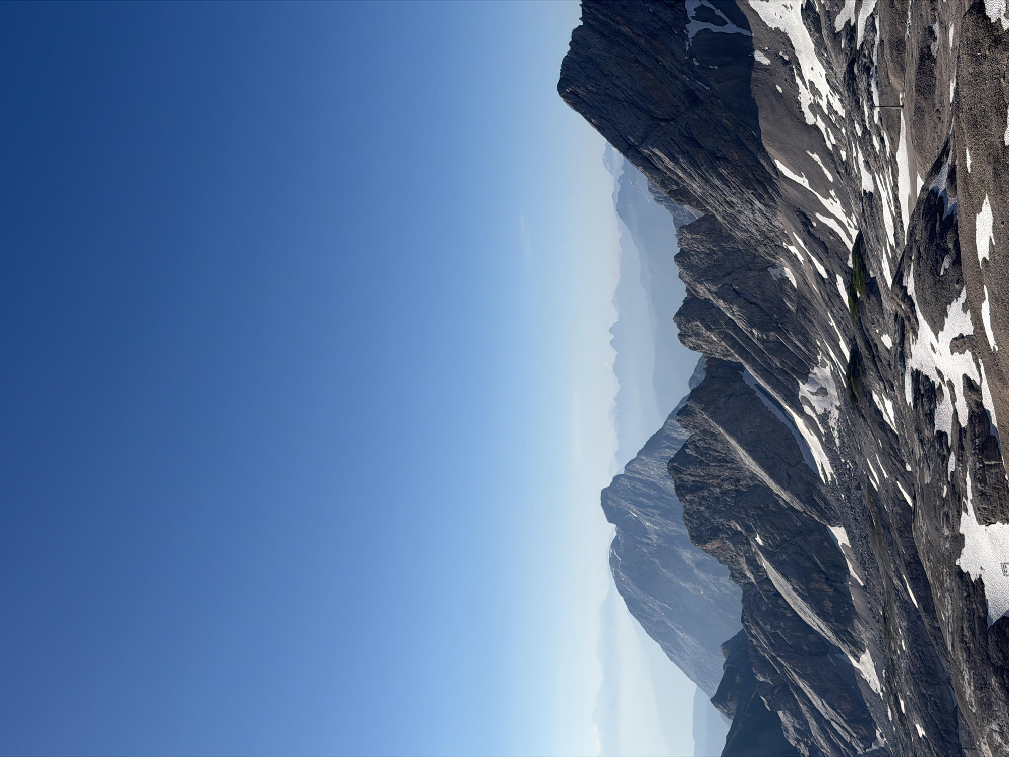 This is an outdoor, vertical, eye-level shot of a rocky mountain range under a clear, bright blue sky. The image is rotated 90 degrees clockwise. The left two-thirds of the image is a vast expanse of clear blue sky. The right third of the image shows rugged, dark gray mountain peaks with patches of white snow scattered across their surfaces. The mountains recede into the distance, appearing lighter and hazier due to atmospheric perspective. The terrain is steep and jagged, with visible layers and fractures in the rock formations. There is no text visible in the image. The lighting suggests it is daytime, likely morning or afternoon, given the brightness of the sky and the clear visibility of the mountain details. The overall impression is one of grandeur and the vastness of a mountain landscape.