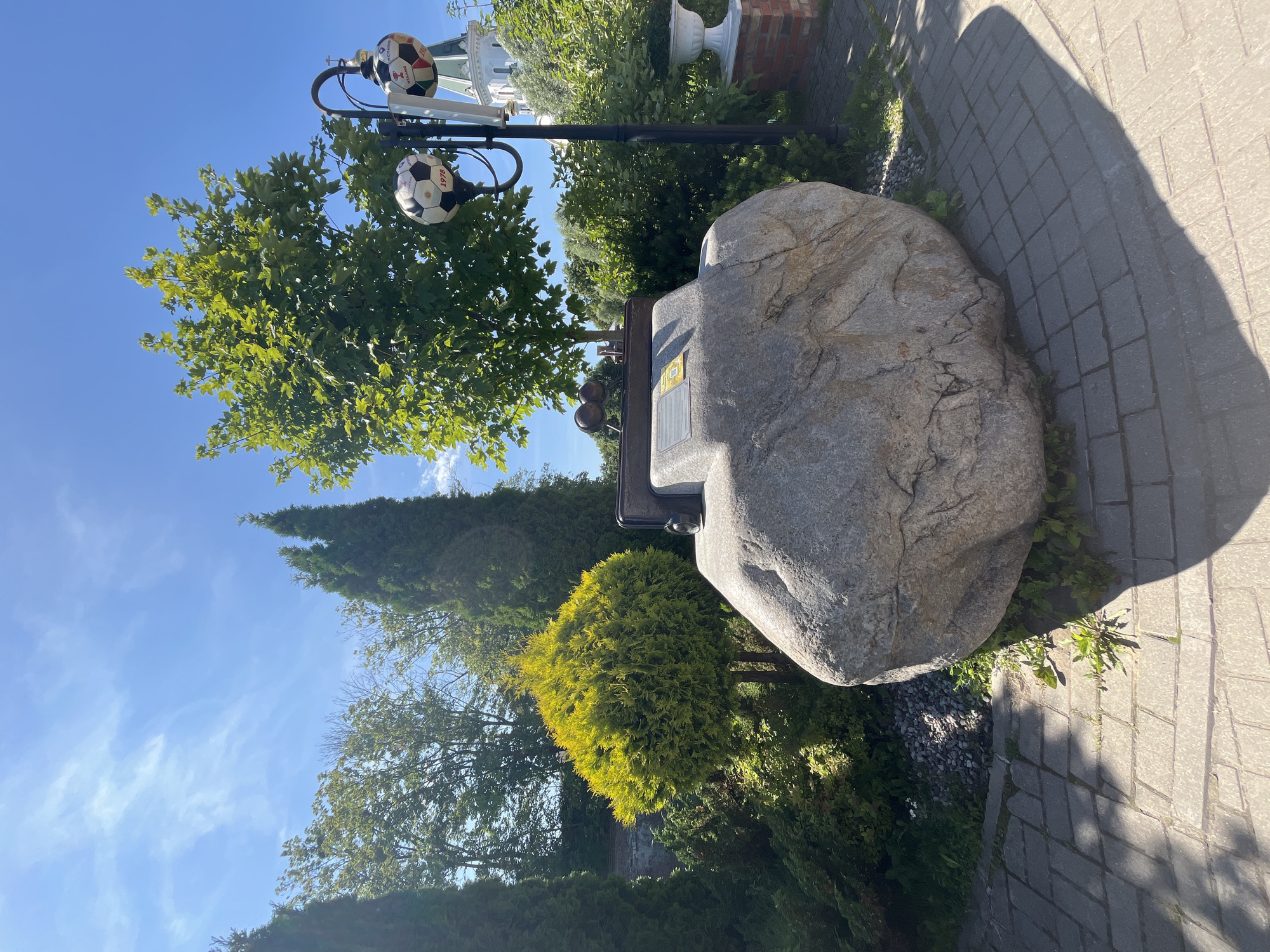 The image depicts an outdoor scene with a large granite boulder as the central subject. Mounted on top of the boulder is a metallic structure with two circular objects, possibly trophies or decorative elements, and a plaque. To the right of the boulder, a lamppost is visible, adorned with two soccer balls, one of which has the number "1978" printed on it. In the background, lush green trees and foliage are abundant, with a building featuring a green steeple and cross visible behind the trees. The ground is paved with interlocking bricks, and the sky is clear blue with scattered white clouds. This memorial is located in Kaliningrad, Russia, specifically in the vicinity of the Church of the Holy Redeemer, which is a landmark in the city. The area appears to be a park or a public square, given the presence of the monument and manicured greenery. No people are present in the image.