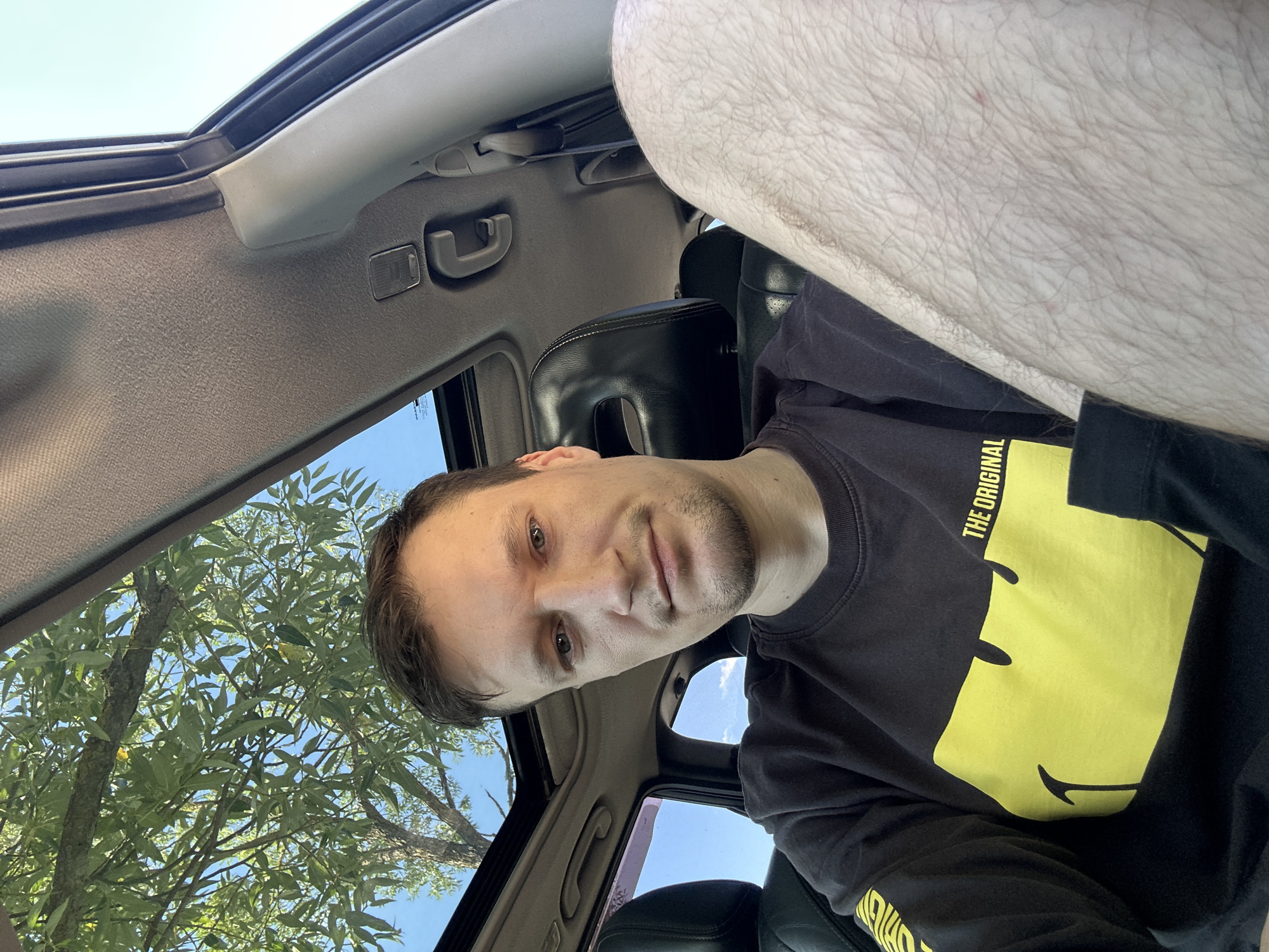 In the city of Makiv, Ukraine, a man is pictured inside a car. The vehicle has a sunroof open, allowing a view of a tree with green leaves and the sky. The man is wearing a black long-sleeved shirt with a yellow graphic design, and the words "THE ORIGINAL" are visible on the sleeve. He is looking towards the camera, with his right leg prominently in the foreground. The interior of the car shows black leather seats and parts of the dashboard and headliner.