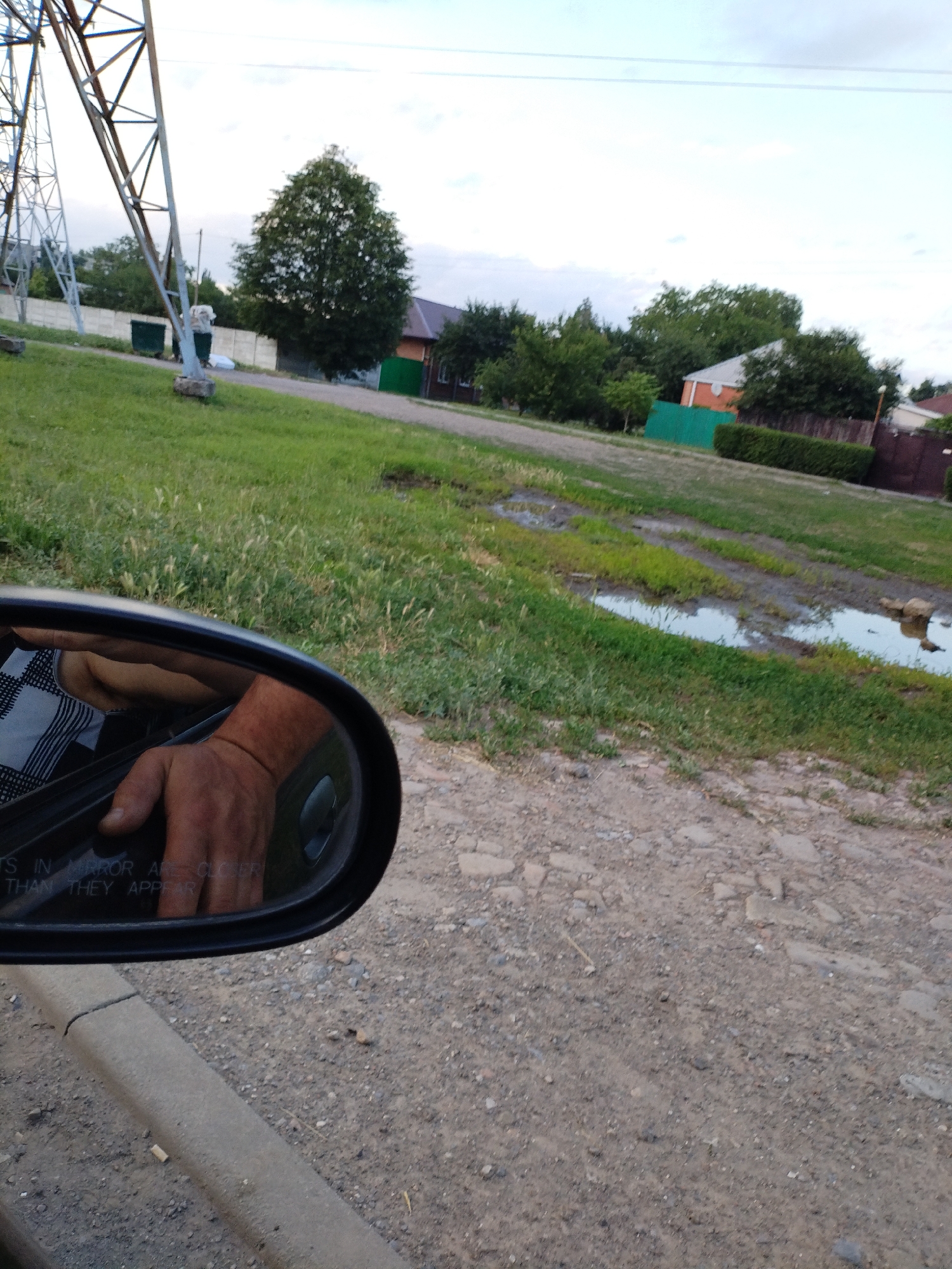 The image depicts a view from inside a vehicle, showing a reflection in the side mirror of a person's arm and hand resting on the car door. The background is a rural or semi-rural scene in Taganrog City District, Russia. A tall metal structure, likely a power line pylon, is visible on the left. Beyond a grassy area with some puddles, there are houses with green and orange accents, as well as fences and trees, suggesting a residential area. The weather appears to be overcast or nearing dusk, with a pale sky. The ground is a mixture of gravel and dirt, with some patches of grass. The text "TS IN MIRROR ARE CLOSER THAN THEY APPEAR" is visible in the lower portion of the side mirror. The overall impression is that of a casual drive through a moderately developed area.