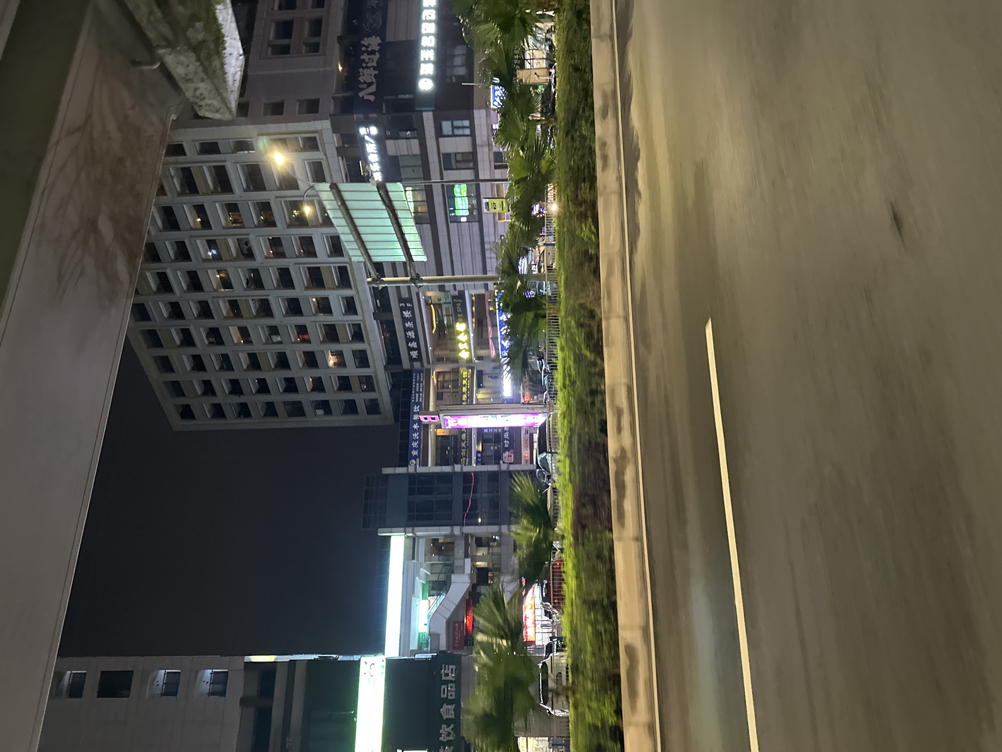 A nighttime street view in Chongqing High-tech Zone, China, captures a busy urban environment. Tall buildings, illuminated by artificial lights, dominate the skyline. One prominent building displays a grid-like pattern of windows, with some windows lit from within. Another building, to the left, is a lighter color and also features multiple floors with windows.
A multi-story commercial building is situated in the mid-ground, featuring a mix of architectural styles. It has a central section with an open-air staircase and escalators, visible through windows. Various signs with Chinese characters and neon lights adorn the facade of this building, indicating businesses like restaurants and shops.
In the foreground, a road with lane markings is visible, suggesting it's a thoroughfare. A median strip planted with lush greenery and palm trees separates the road from the sidewalk and buildings. Several cars are parked along the sidewalk or are visible on the road. The overall scene depicts a vibrant and active city at night, with commercial activity and transportation in full swing.