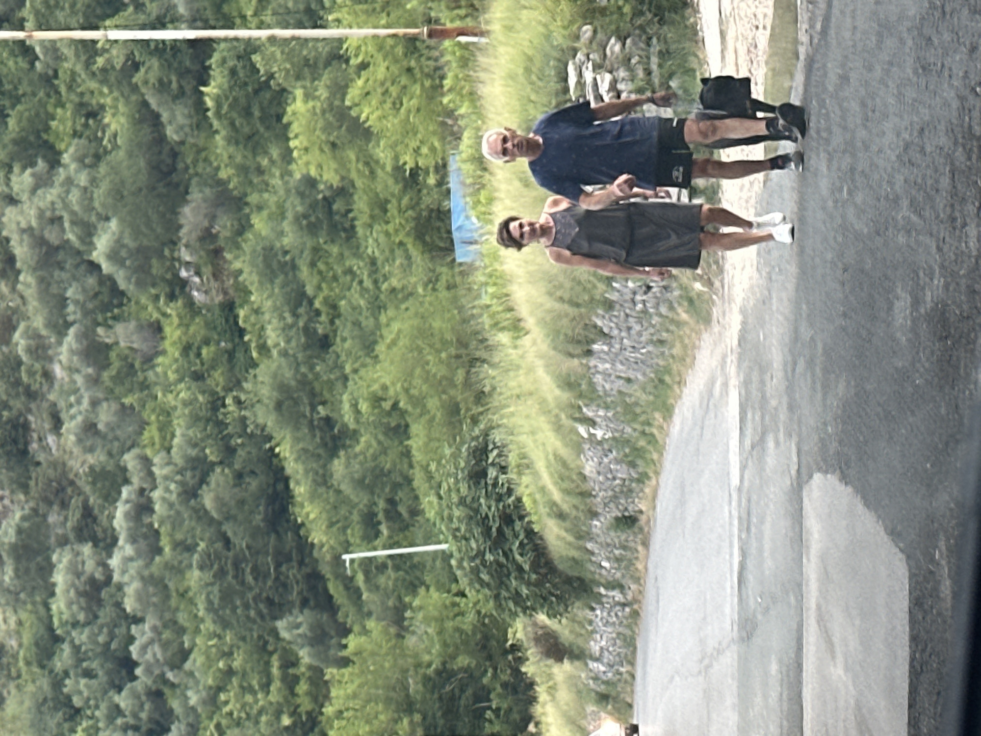 Two people are walking on a paved road in a hilly, green landscape. The person in front is wearing a dark-colored dress and white sneakers. The person behind them is wearing a blue t-shirt, dark shorts, and black athletic shoes. A black bag is visible near the feet of the person in the blue t-shirt. The scene appears to be outdoors during the daytime, with lush green foliage covering the hillsides. The road is asphalt and wet in places, suggesting recent rain or a damp environment. The presence of trees and the mountainous terrain indicate a natural, possibly rural or semi-rural setting. The people's posture and their direction of movement suggest they are engaged in a walk or a leisurely stroll. There is no visible text in the image.