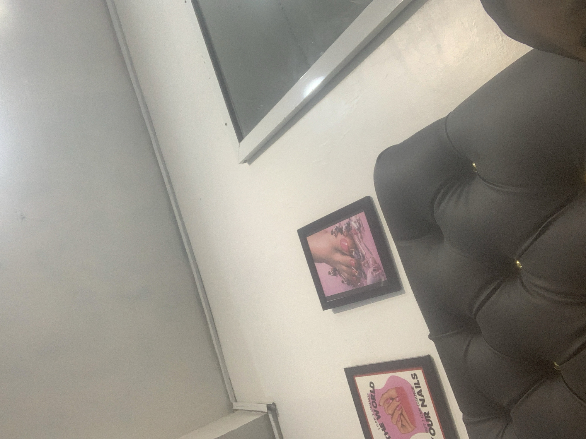 A framed artwork featuring a woman's hands with manicured nails and the text "BUT I CAN'T CHANGE THE WORLD" is displayed on a white wall. Below it, another framed artwork depicts a close-up of feet with polished toenails and decorative elements, possibly flowers. The artworks are positioned above a dark upholstered furniture piece, which appears to be a chair or sofa. To the right of the artworks is a white wall that continues to the upper right corner of the image. The scene is consistent with the interior of a nail salon or a similar establishment in Moba, Nigeria.