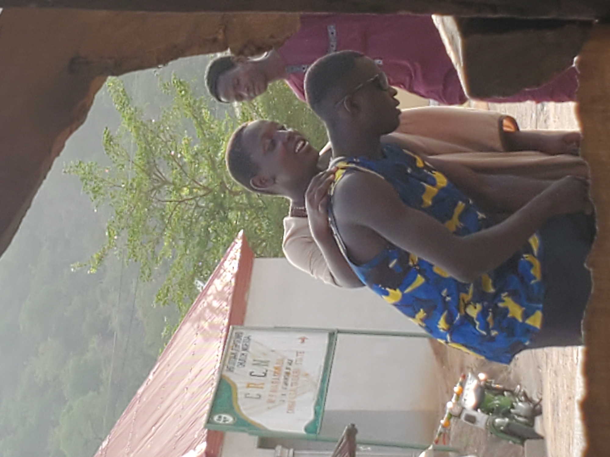 The image captures a casual outdoor scene featuring three young males, likely adolescents or young adults, against a backdrop of lush greenery and distant hills. The perspective is from inside a structure, looking outwards through an archway or opening, with parts of the frame visible at the top and right.
In the immediate foreground, a young man wearing a blue sleeveless top with yellow patterns and sunglasses is seen looking towards the right side of the frame, his mouth slightly open as if speaking or observing something intently. Behind him, another young man in a light-colored t-shirt has his arm casually around the first's shoulder, smiling broadly or laughing with an open mouth, conveying a sense of camaraderie and joy. Further back, a third young man, dressed in a magenta or purple long-sleeved shirt, looks directly at the camera with a subtle, friendly smile.
To the left of the subjects, a building with a reddish-brown corrugated metal roof and white walls is visible. Attached to this building is a rectangular sign with a green and gold decorative border. Prominently displayed on the sign are the letters "E.R.C.N." with "MARARABA" written below it. This text strongly indicates the location is Mararaba, Nigeria, and suggests the building is an establishment of the Evangelical Reformed Church of Christ Nigeria. On the ground near the sign, some small household items or containers are scattered, including what appears to be a small kettle.
The scene is bathed in bright daylight, suggesting a clear or possibly hazy day. The vibrant green foliage underscores a natural, perhaps humid, environment. The overall mood is relaxed and friendly, with the individuals appearing engaged in conversation or enjoying each other's company.