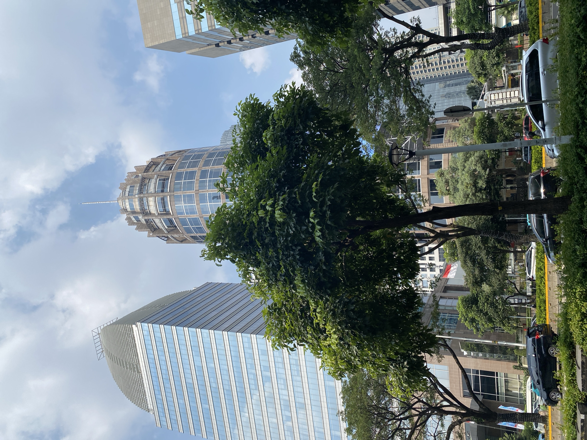This image shows a street scene in Central Jakarta, Indonesia, with tall modern buildings and lush green trees lining the road. The sky is blue with scattered white clouds. In the foreground, a black car is driving on the left side of the road, while a silver car is in the right lane. Several other vehicles are visible in the background, as well as a building with an Indonesian flag flying from it. The buildings are a mix of glass and concrete, with some featuring unique architectural designs. Trees with dense green foliage are strategically planted along the street, providing a contrast to the urban landscape. The overall impression is one of a bustling city with a touch of nature.