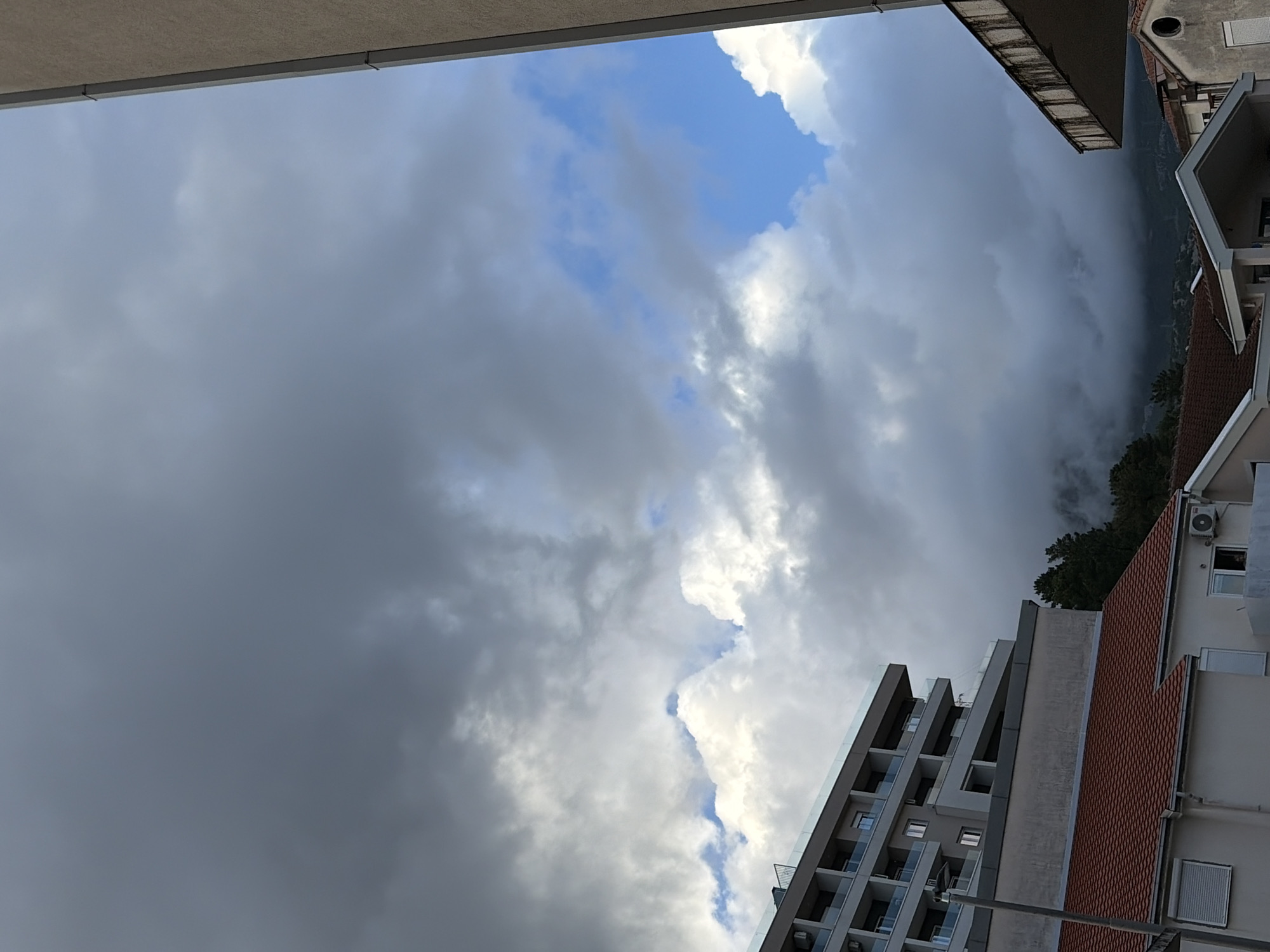 The scene is an upward shot framed by the edges of buildings, likely taken from a balcony in Budva, Montenegro. The sky is filled with dark, stormy clouds, except for a small patch of bright blue. A modern apartment building with multiple balconies and windows is on the lower right, next to a traditional building with a red tile roof. Trees are visible between the two buildings, growing on a hillside.